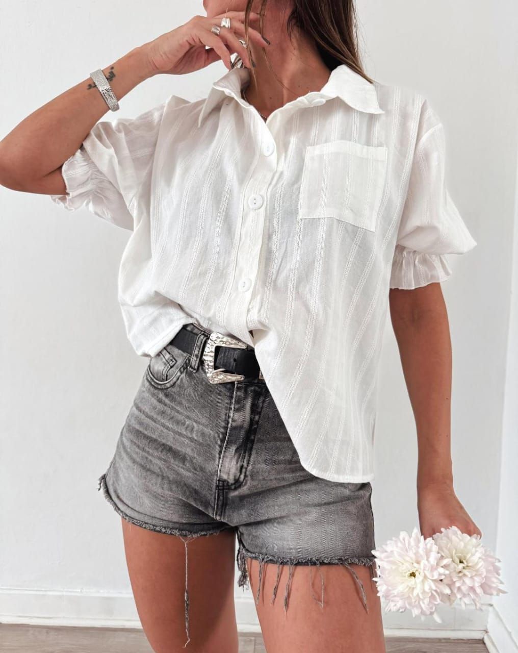 Mujer con camisa blanca y pantalones cortos grises, sosteniendo flores.