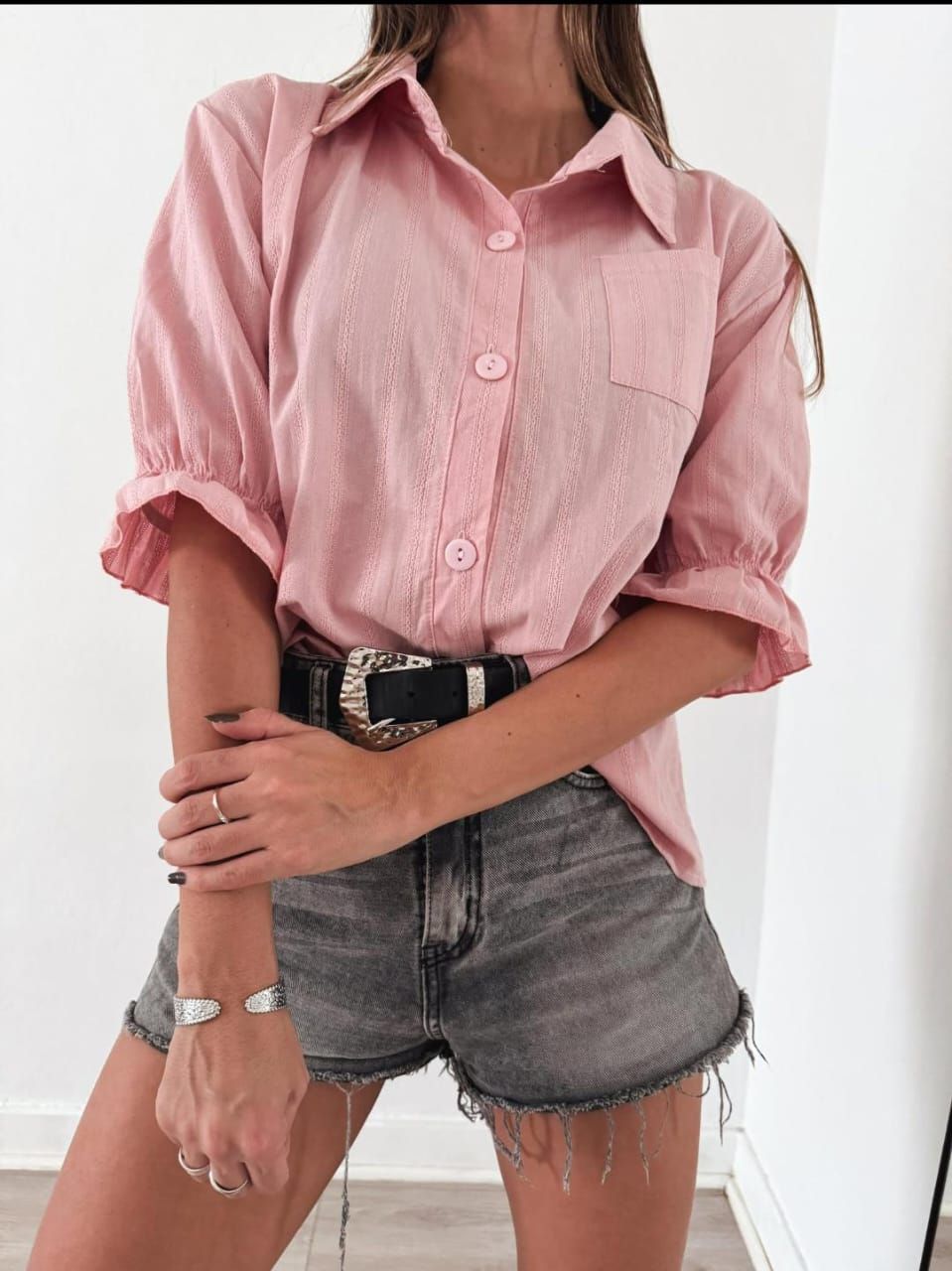 Mujer con camisa rosa, pantalones cortos grises y cinturón, posando en el interior.