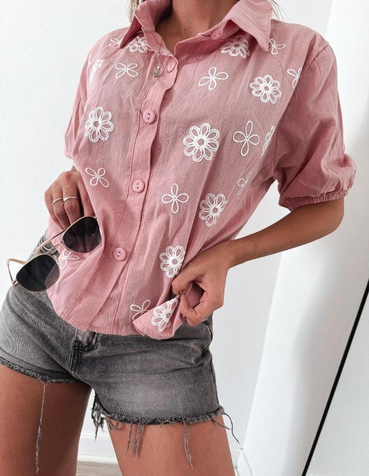 Mujer con camisa rosa bordada con flores y pantalones cortos de mezclilla, gafas de sol metidas dentro, posando casualmente.