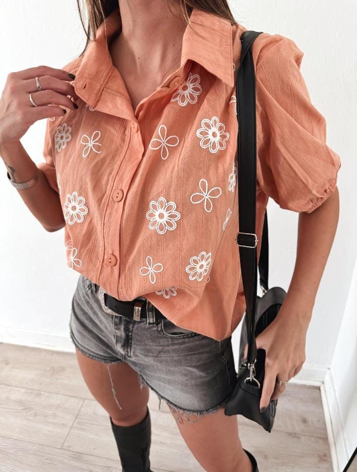 Mujer con camisa bordada en color melocotón y pantalones cortos de mezclilla, con un bolso bandolera negro y botas negras.