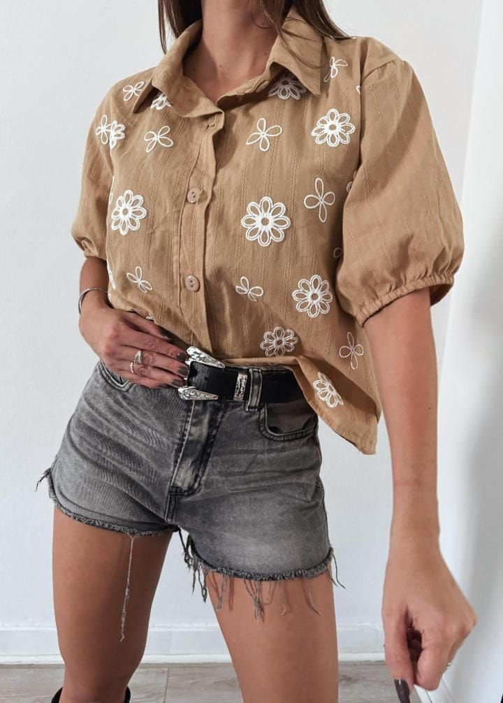 Mujer que lleva una camisa color canela con bordados florales y pantalones cortos de mezclilla grises, de pie en el interior.