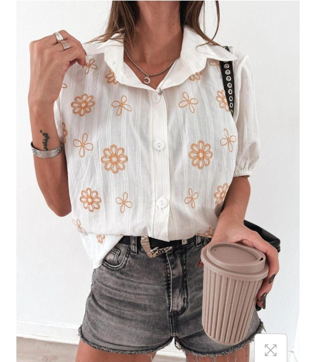 Mujer con una camisa blanca bordada y pantalones cortos de mezclilla, sosteniendo una taza de café.