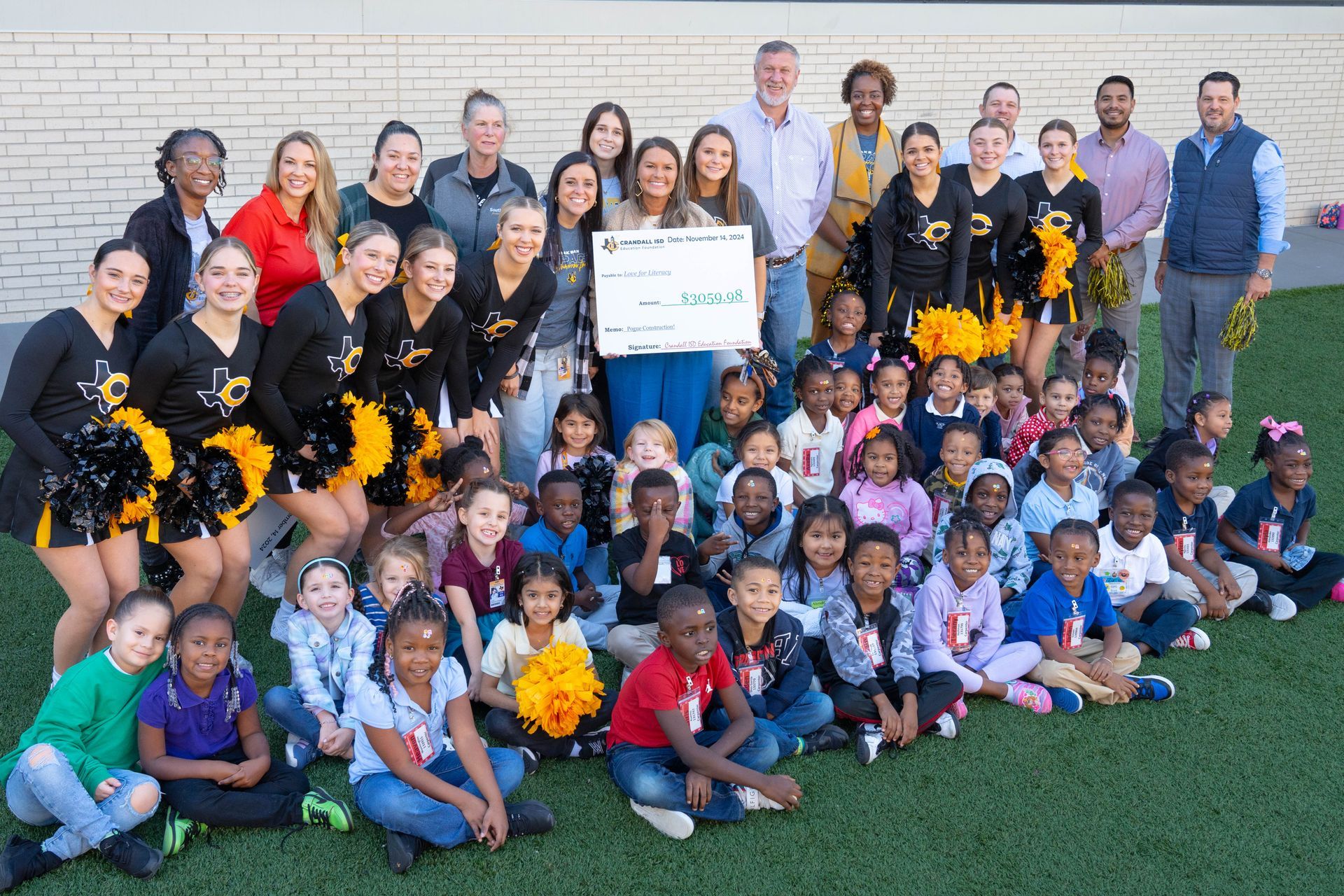 Group photo: Cheerleaders, children, and adults holding a donation check on a green lawn.