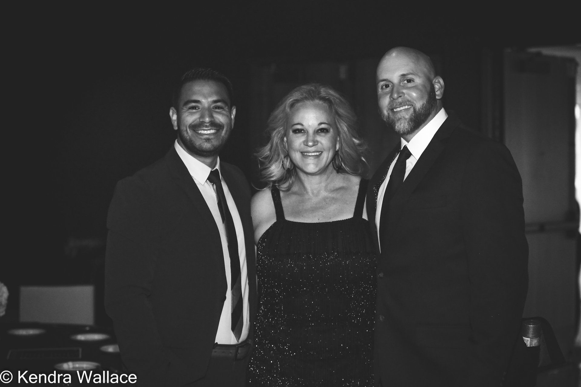 Three people smiling at an event. Woman in a sparkly dress, flanked by men in suits. Black and white.