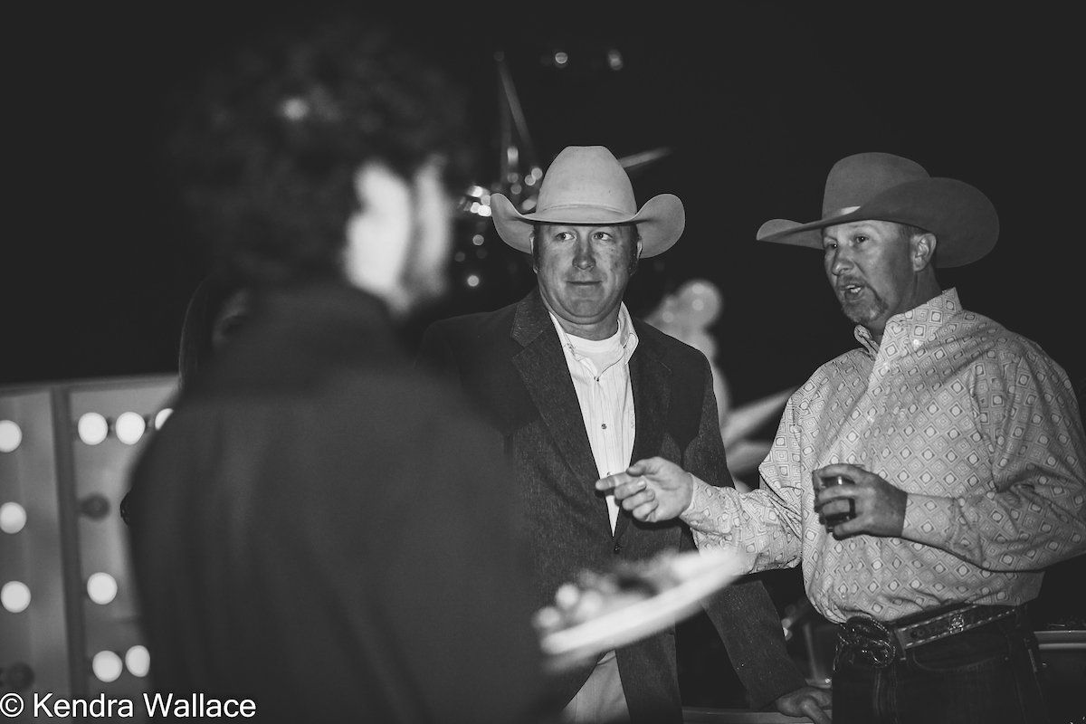 Three men at a nighttime event, two wearing cowboy hats, one pointing, all looking at each other.