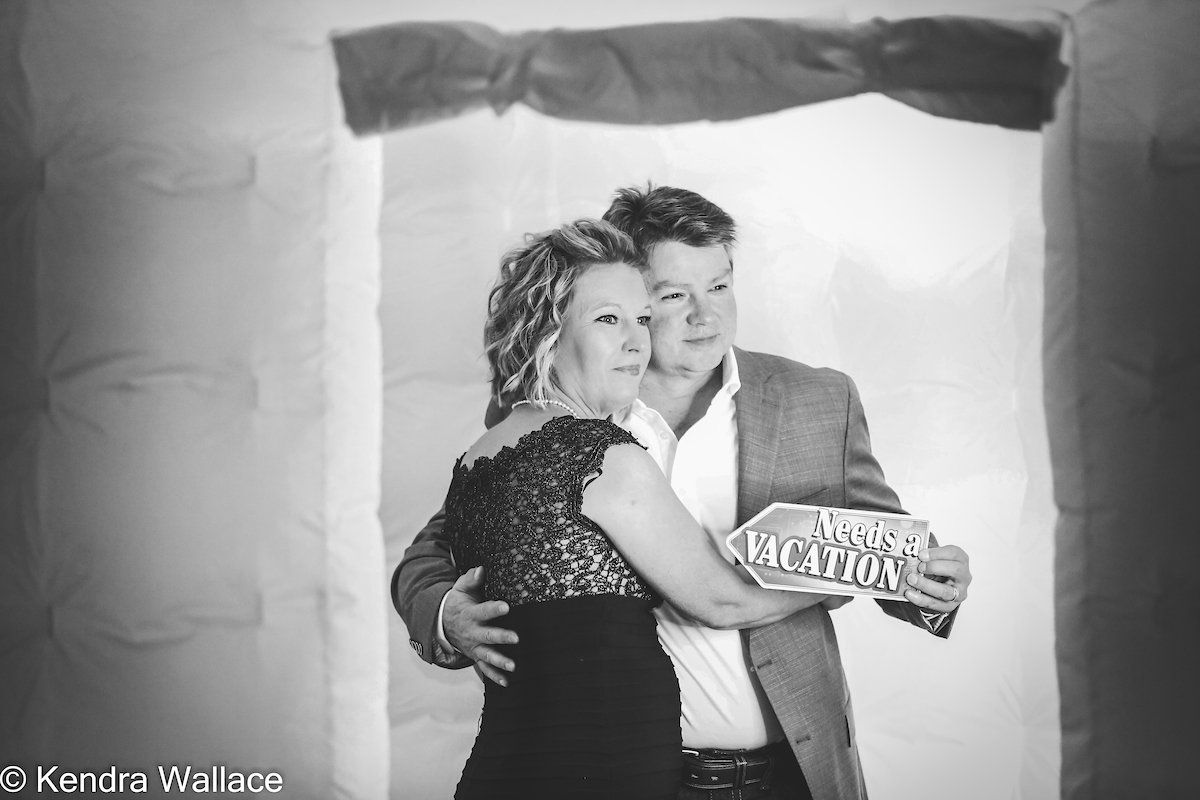 Couple embracing in a photo booth; woman in formal dress, man in blazer, holding a sign.