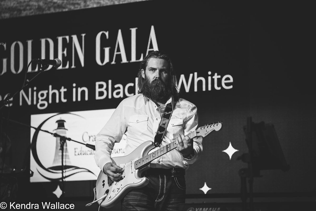 Man with a long beard plays electric guitar on stage at a gala event. 
