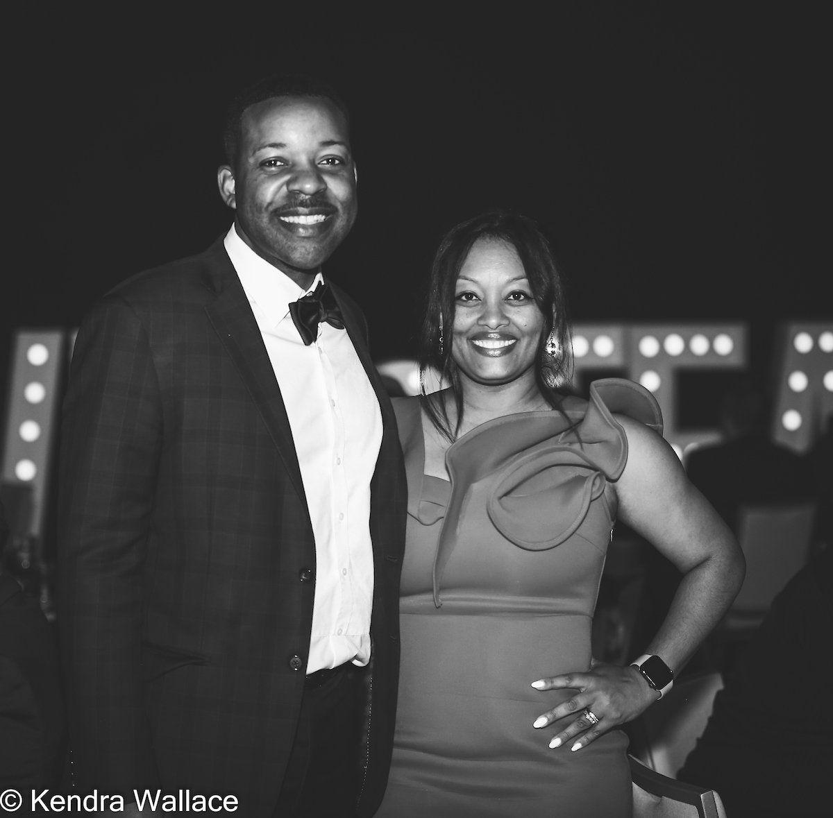 Man in suit and woman in dress pose smiling; lit backdrop, event setting.