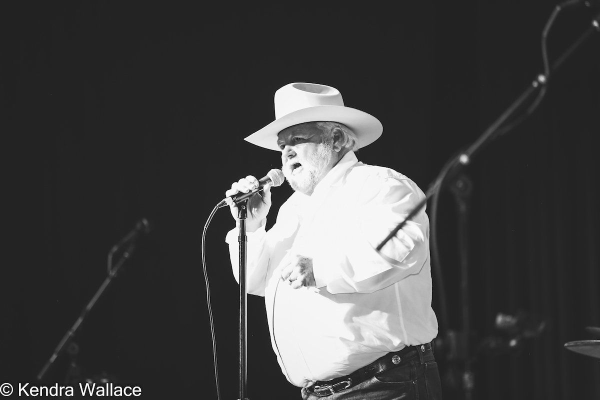 Man in white shirt and hat singing into microphone on stage.