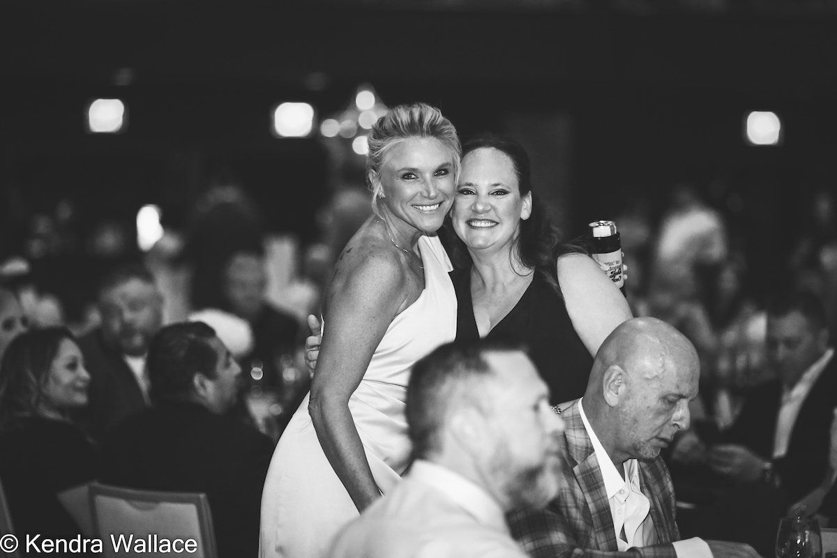 Two women embrace at a formal event. The women smile, surrounded by seated people in a well-lit room.