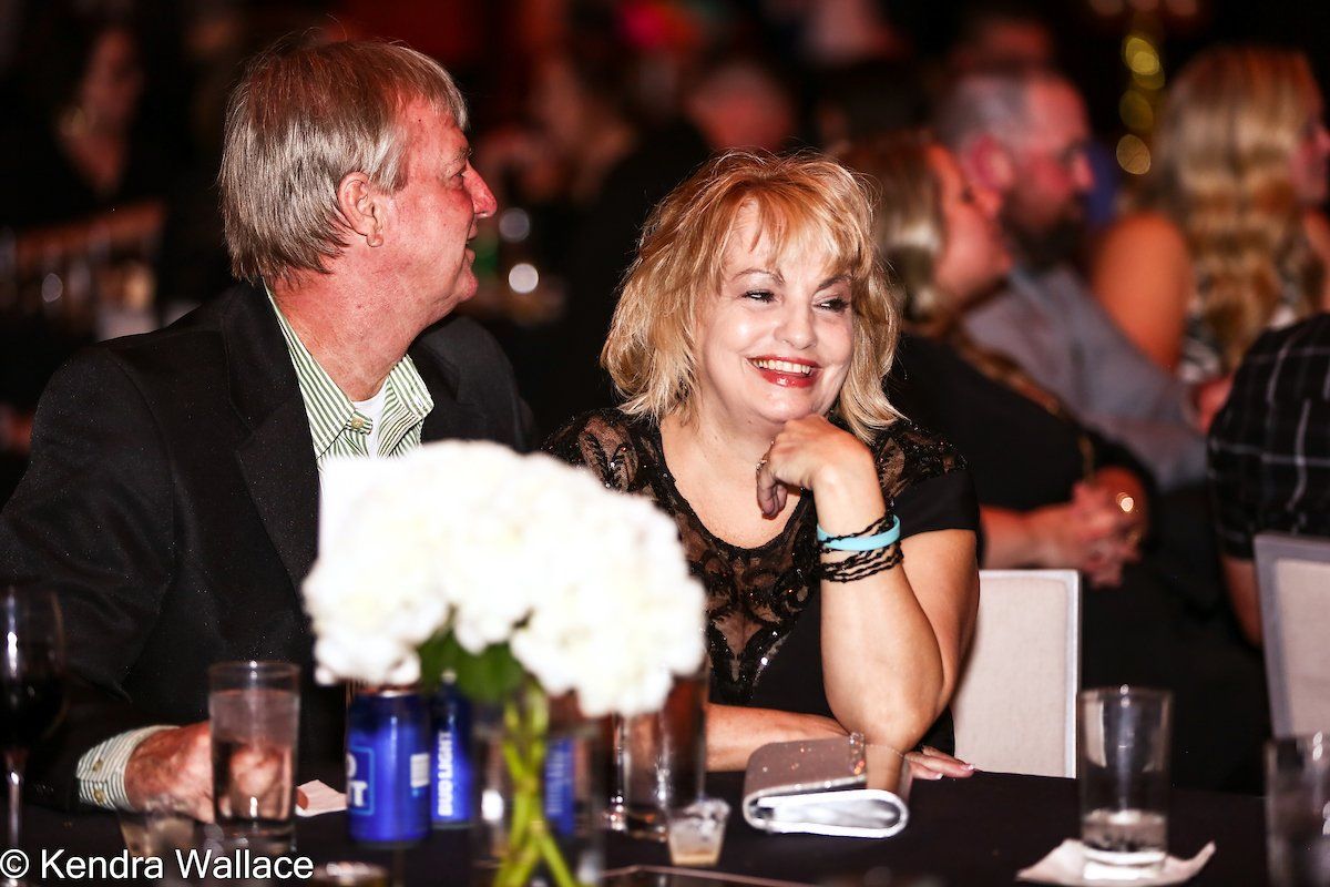 Couple at a gala. Woman smiles, resting chin on hand, wearing black lace. Man looks on, wearing a blazer.