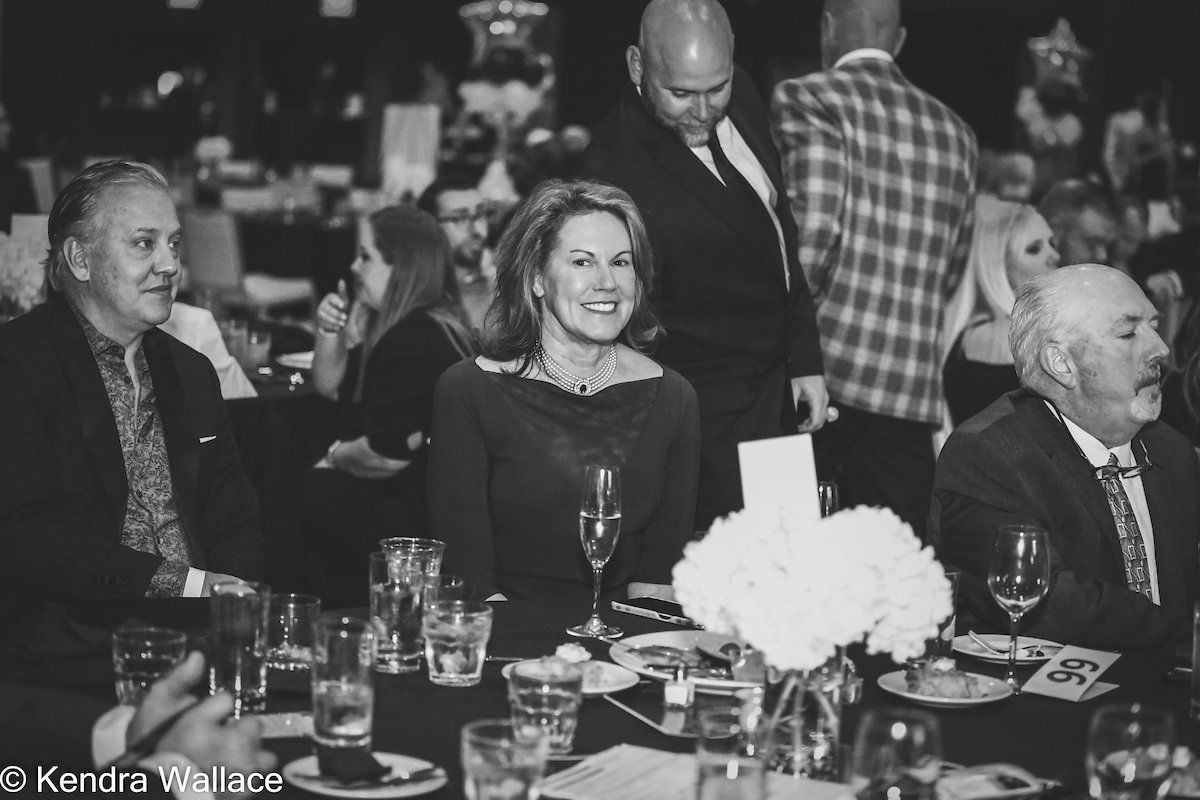 People at a formal event seated at a table. Woman smiles, others have varying expressions. Black and white.