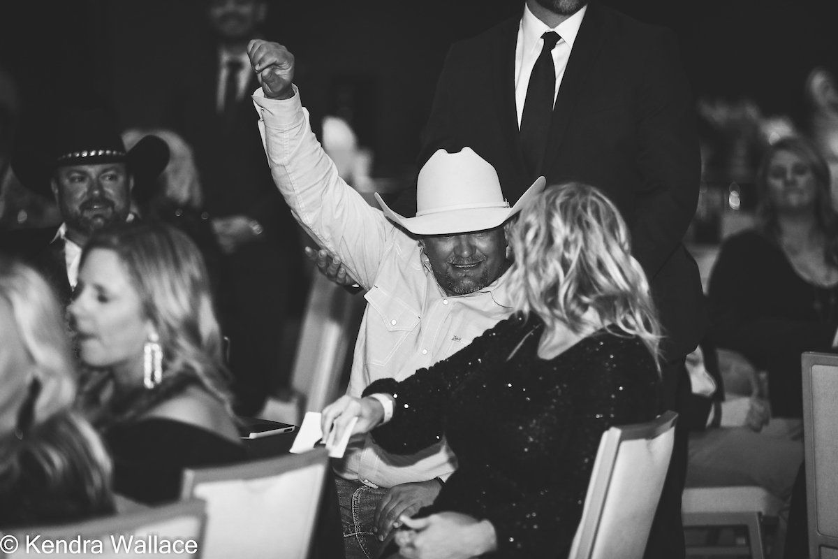 Man in cowboy hat raises arm, with a woman in a black dress. Event setting, people in the background.