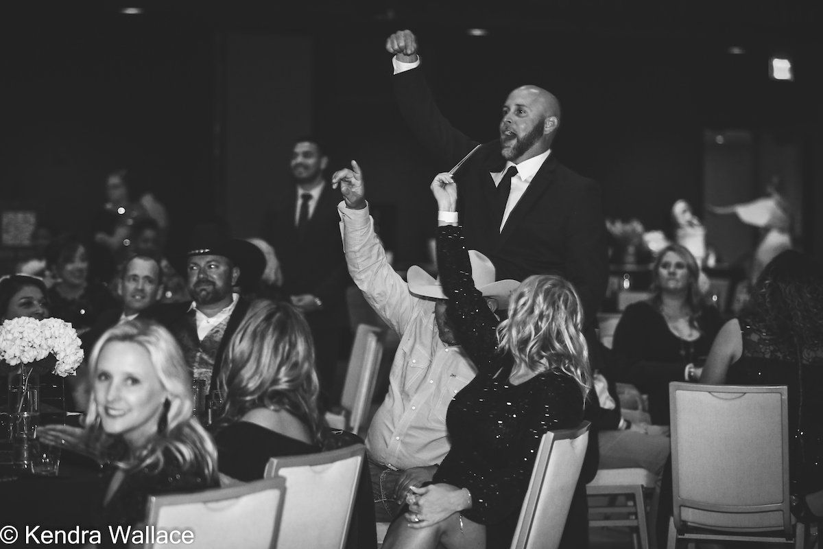 People at a formal event raise hands during an auction; a man in a suit gestures from a raised platform.