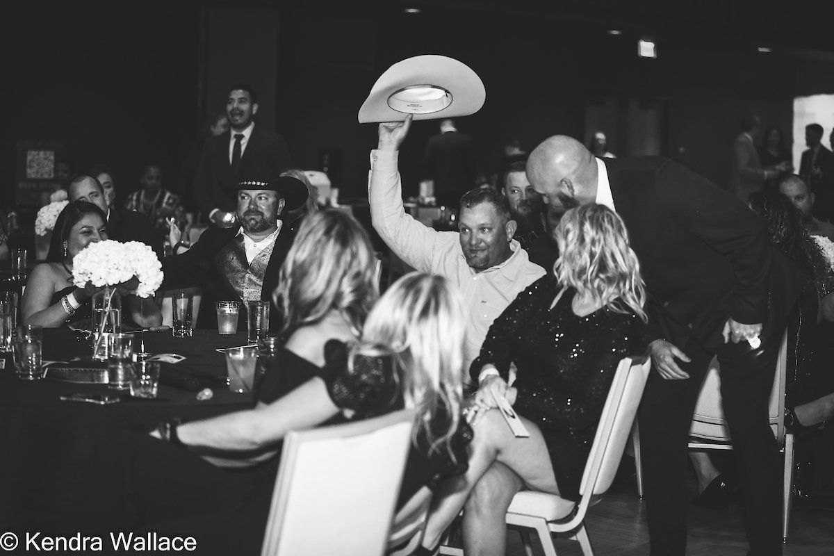 Man raising cowboy hat at a formal event. Guests seated at tables. Black and white.
