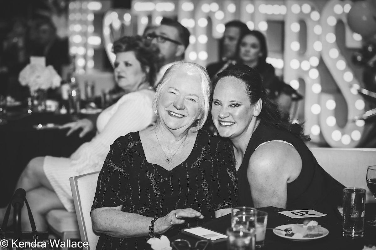 Two smiling women at a formal event, seated near a table. Backlit letters in background. Black and white.