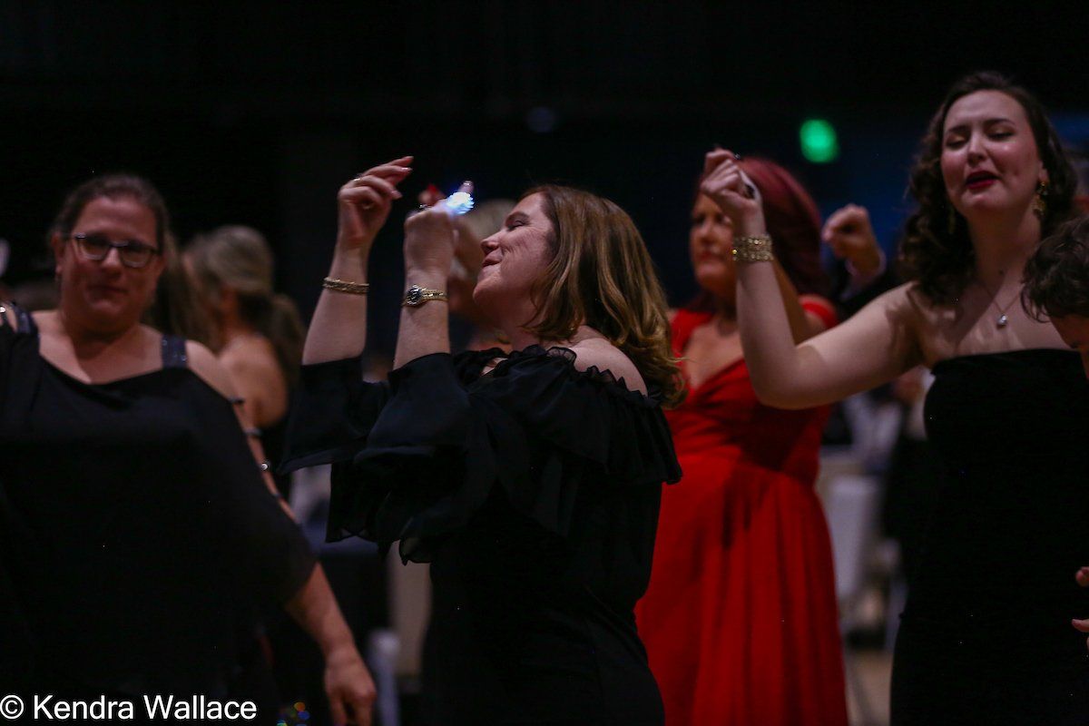 People dancing at an event, woman in black dress raising arms, other women nearby.