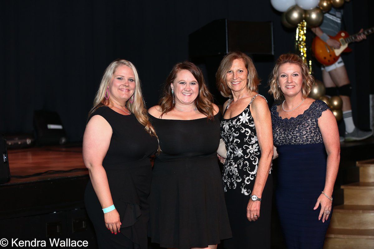 Four women in formal wear pose together on a stage with balloons and a guitar player visible in the background.