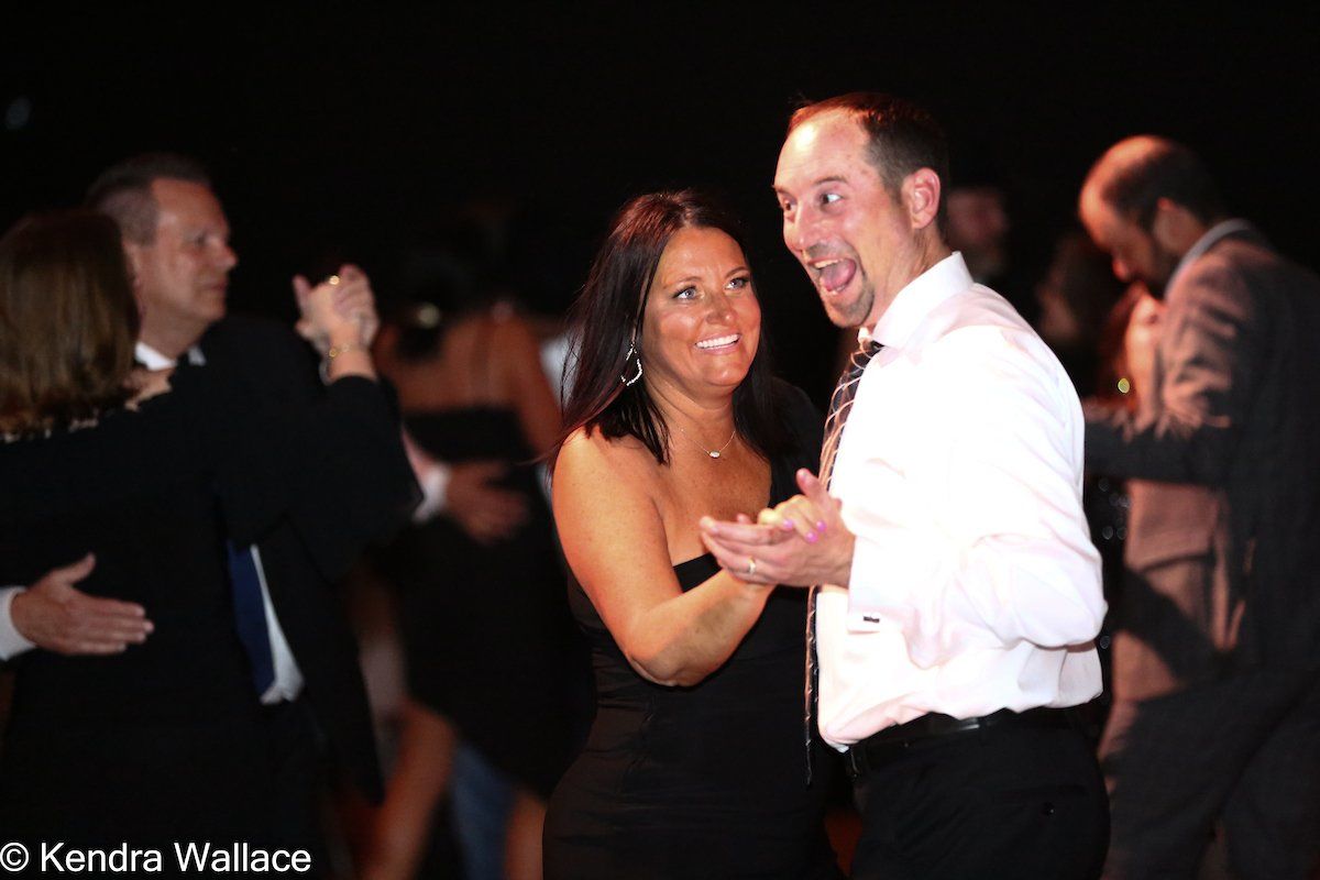 Couple dancing at an event; woman in black dress smiles, man in white shirt laughs. Dark background with other dancers.