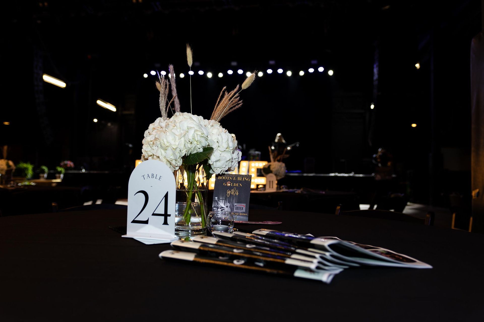 Table setting with floral centerpiece, table number 24, and event programs on a black tablecloth. Stage in background.