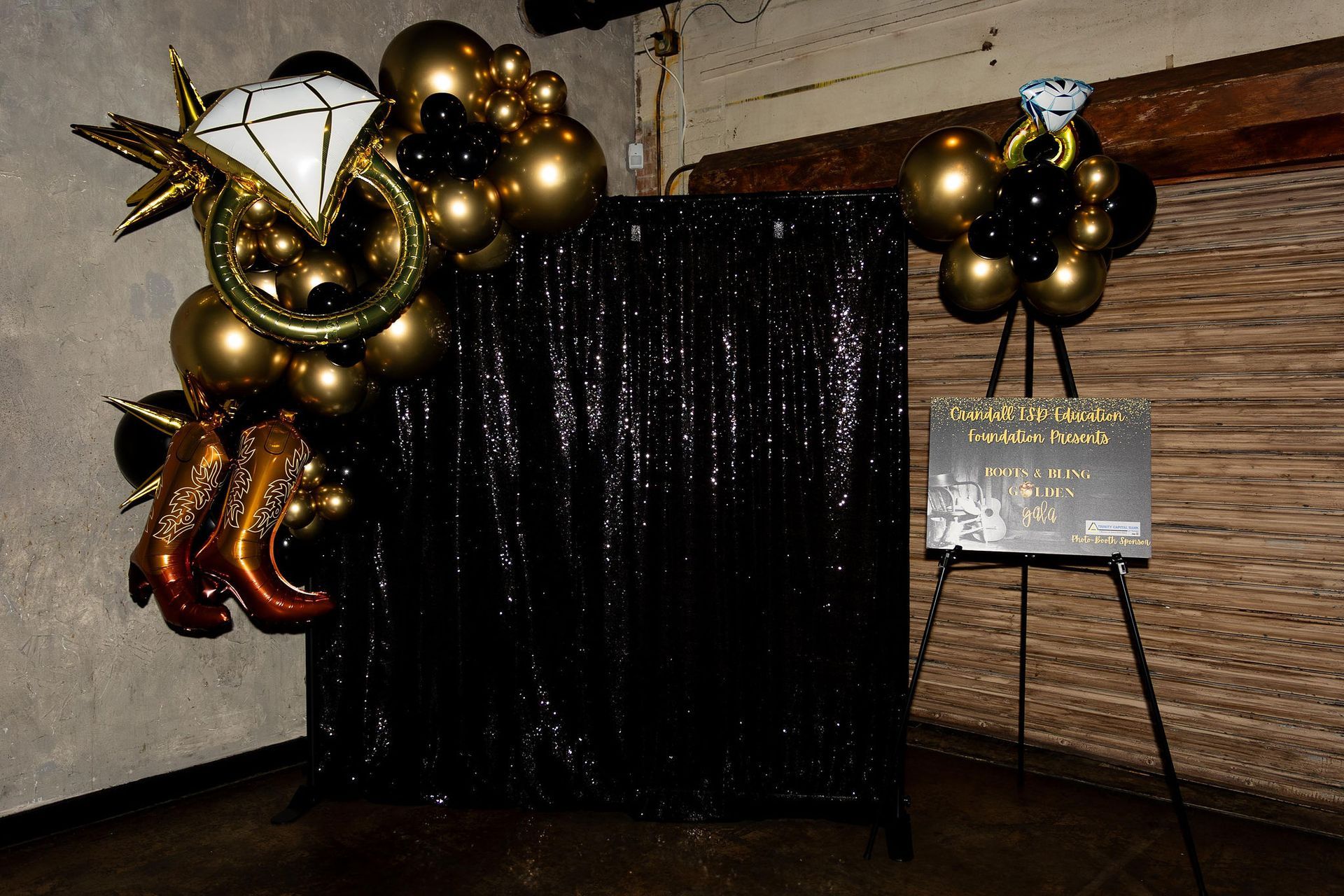 Black sequin backdrop with gold and black balloon decorations for an event; sign on an easel.