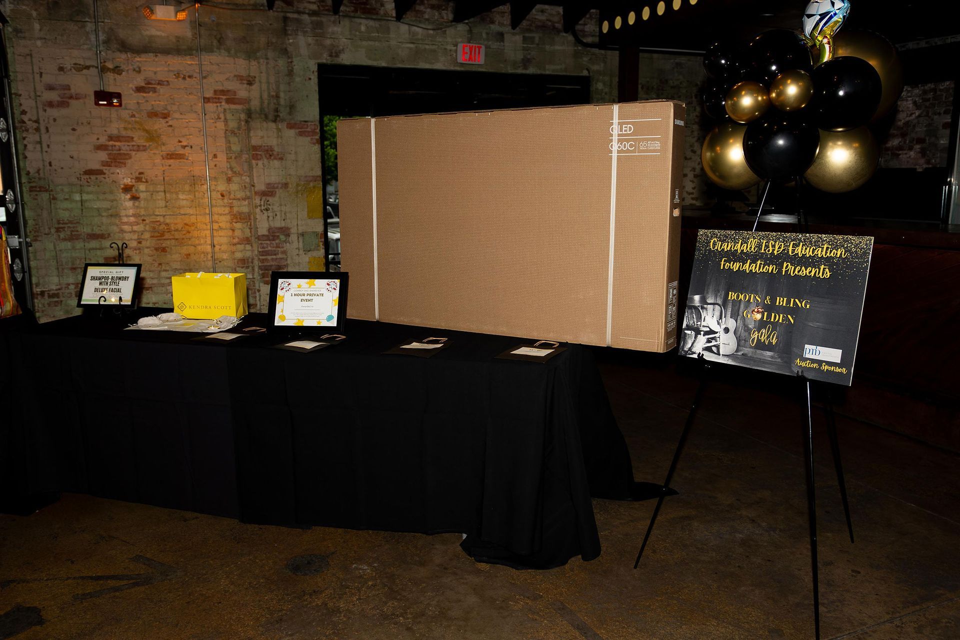 Black-draped table with awards, cardboard box, and chalkboard sign; balloons and brick wall in background.