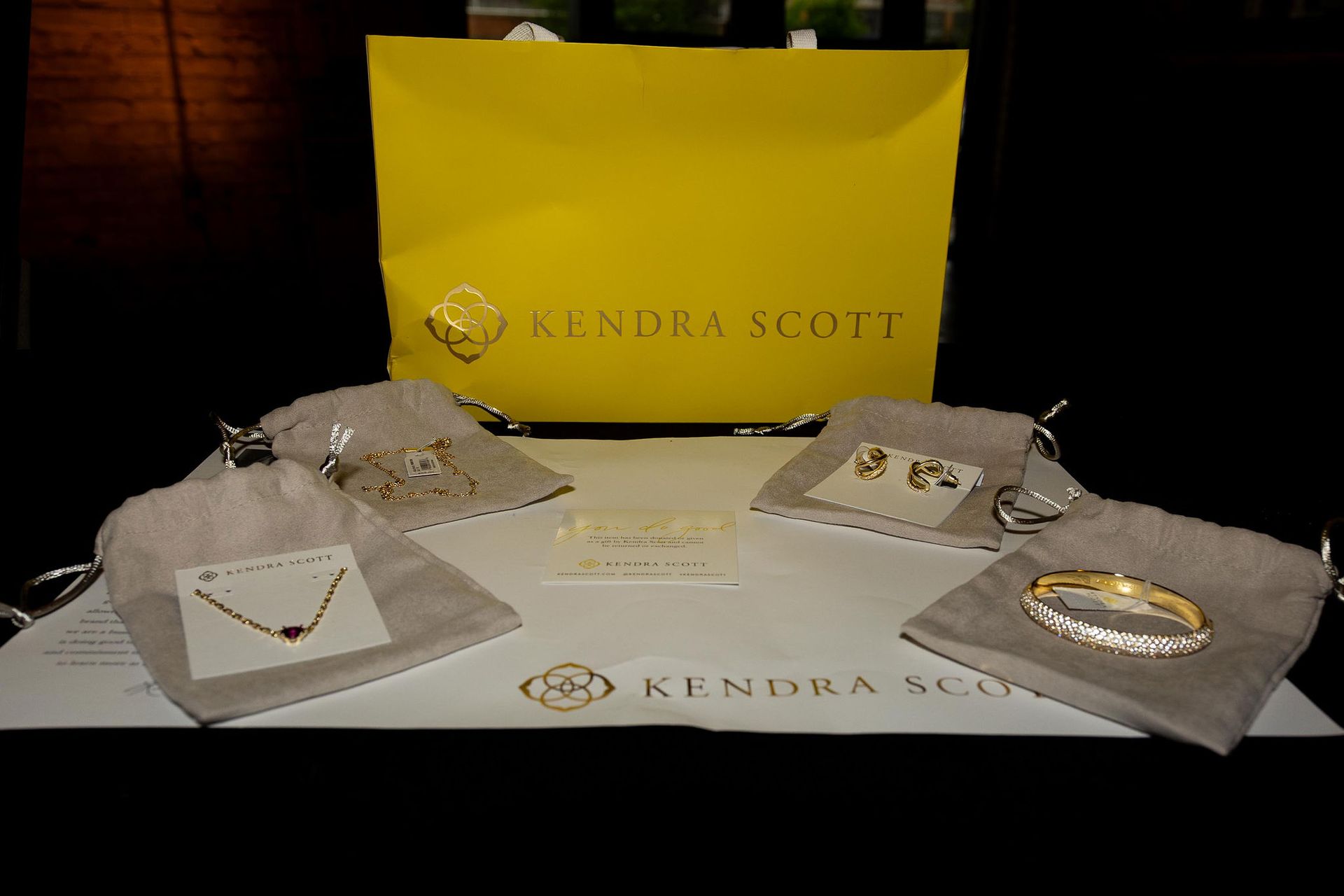 Kendra Scott jewelry display with a yellow bag, small gray bags, and gold necklaces, earrings, and a bracelet.