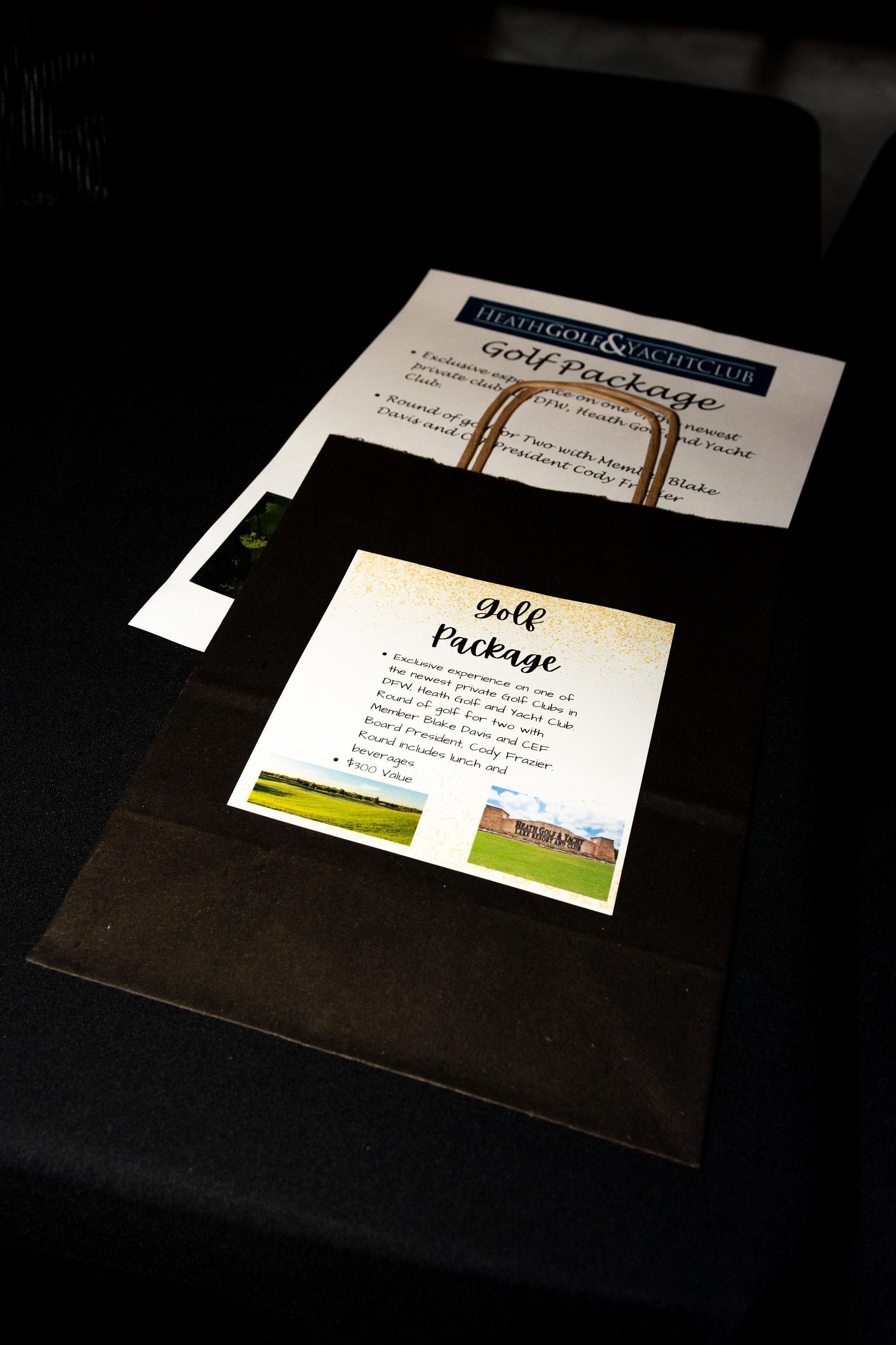 A black paper bag with a golf package flyer and a smaller flyer on a dark surface.