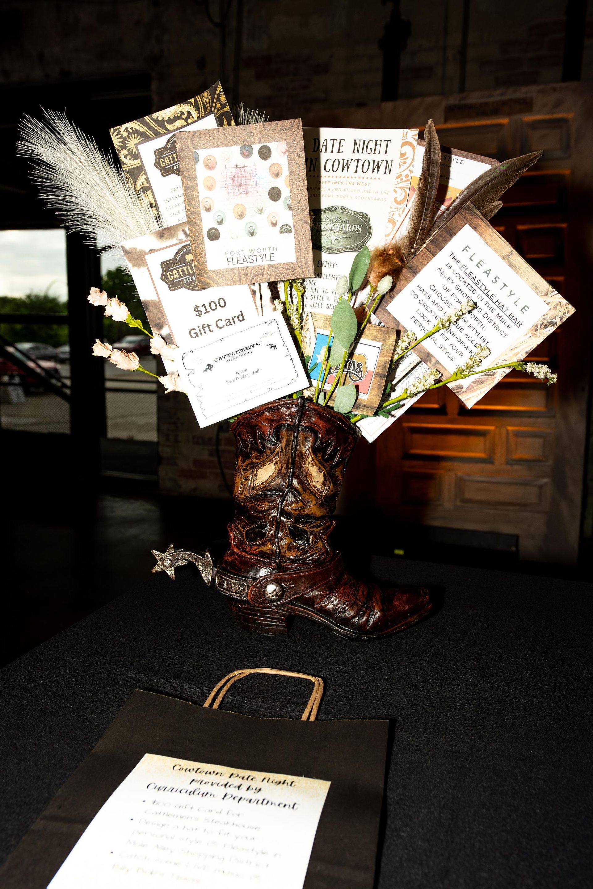Cowboy boot vase with cards and feathers on a table next to a black gift bag.
