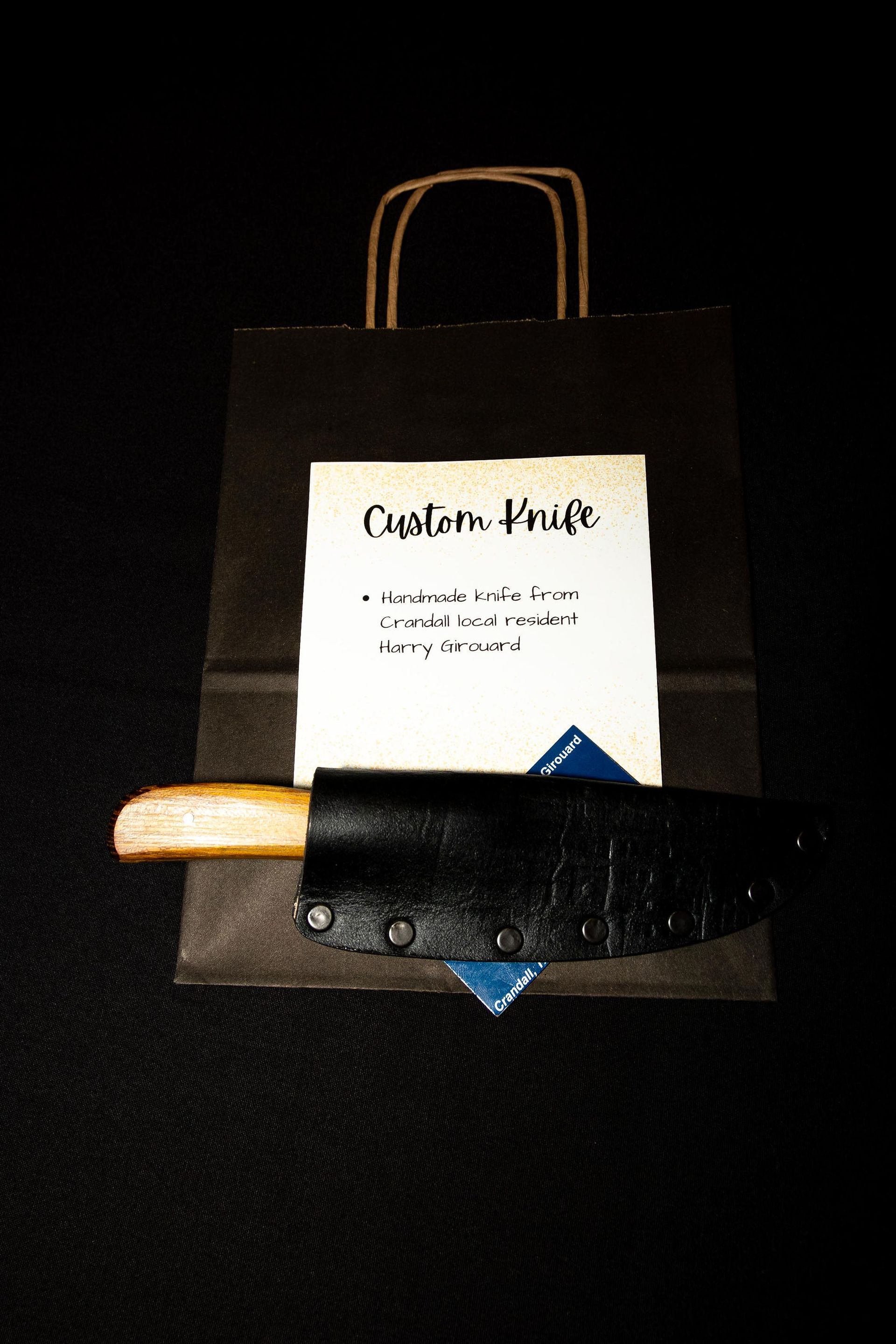 Knife with wooden handle in black leather sheath, leaning against a brown paper bag with a white label, dark background.