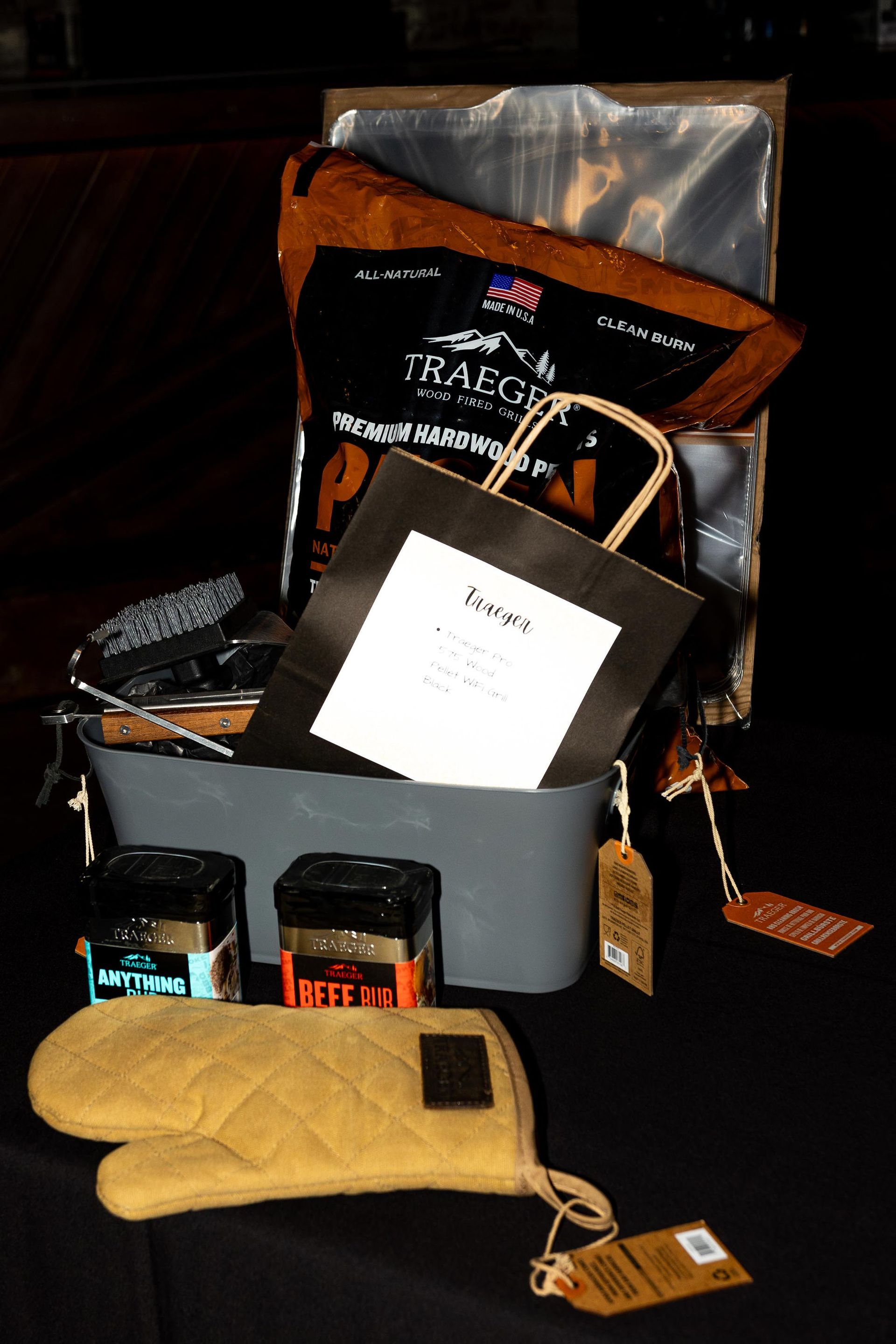 BBQ gift basket with wood pellets, seasonings, tools, oven mitt, and a tote bag on a black surface.