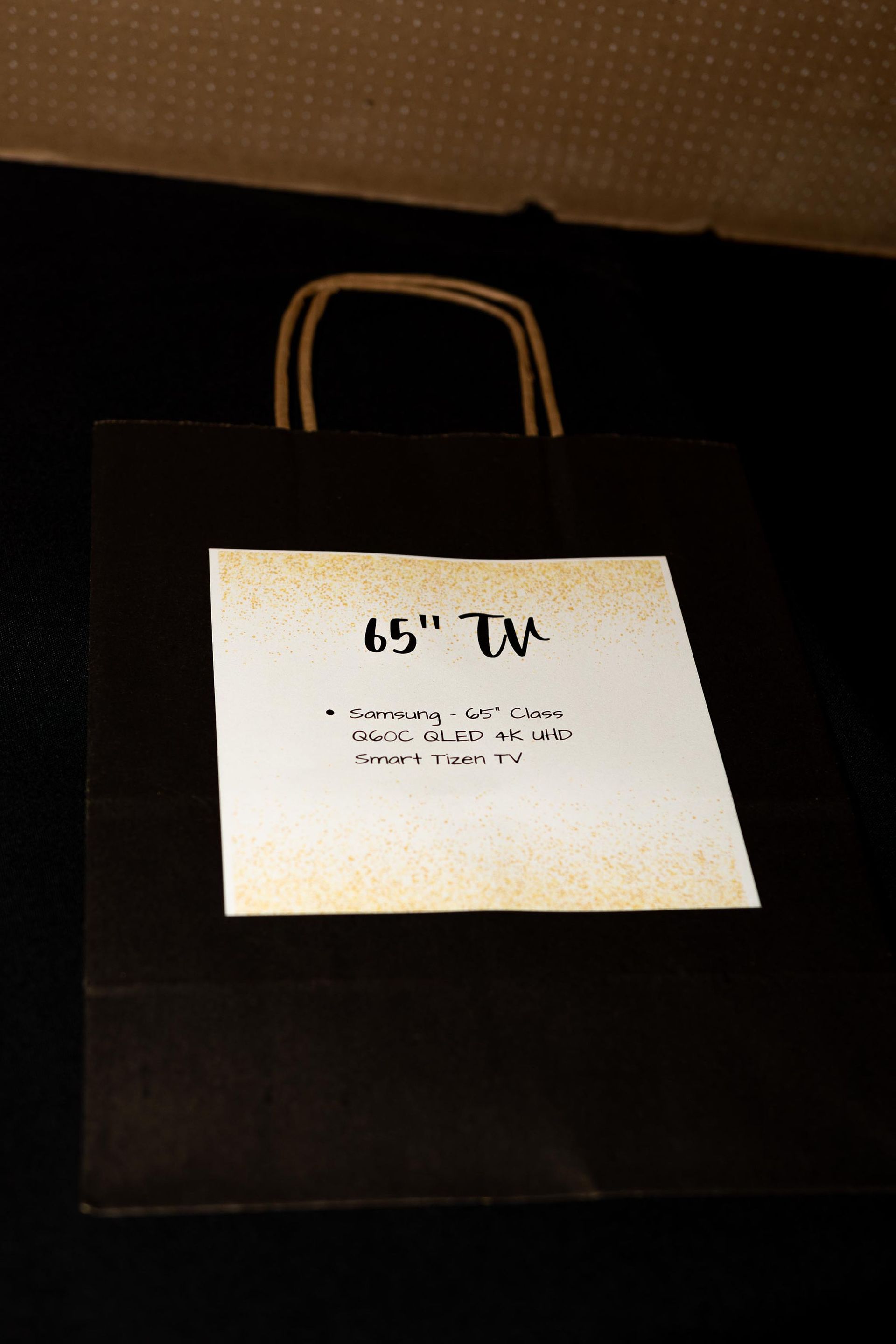 Black gift bag with label stating “65” TV.” Brown handles.
