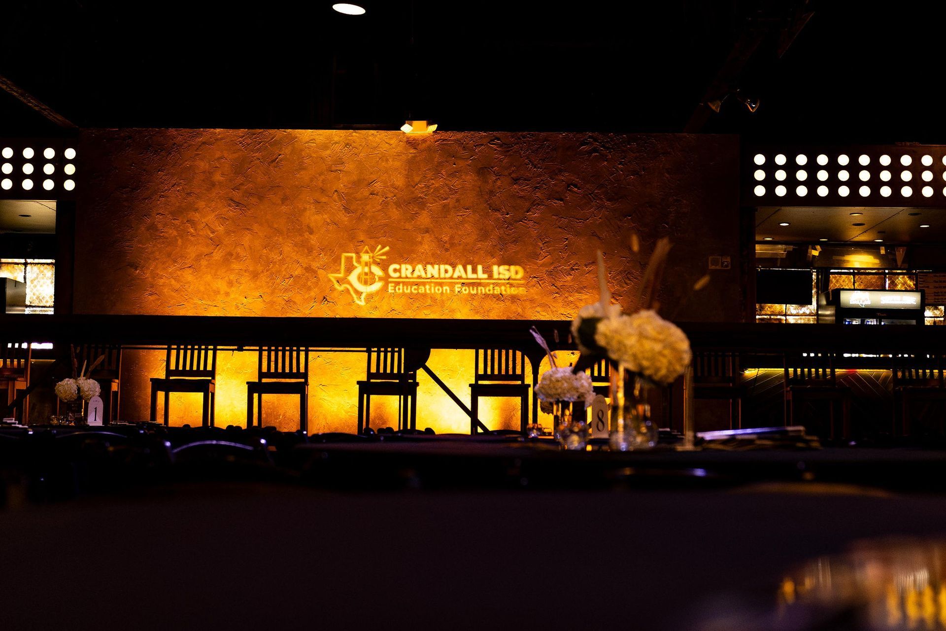 A stage with a golden backdrop and logo for Grand Ball, with chairs and floral decorations.