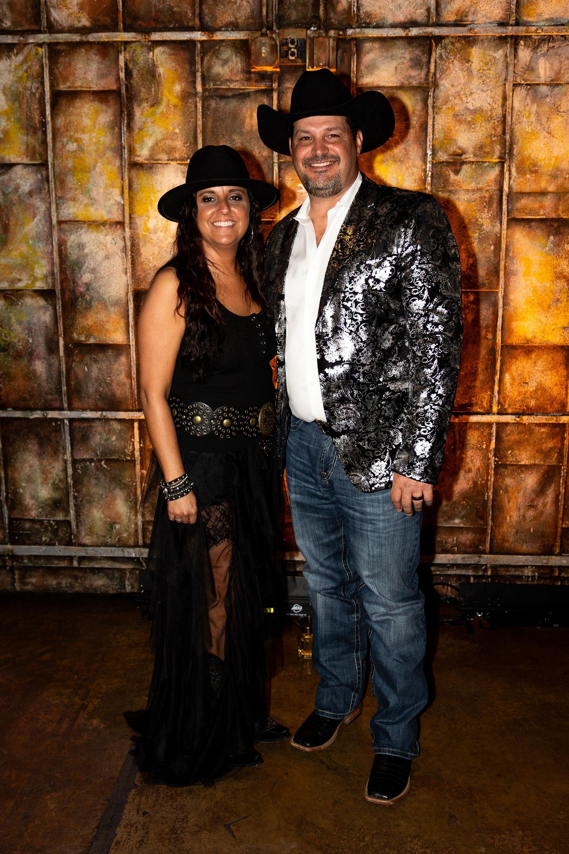 Couple in cowboy hats, posing. Man wears a sequined jacket and jeans; woman in a black dress.