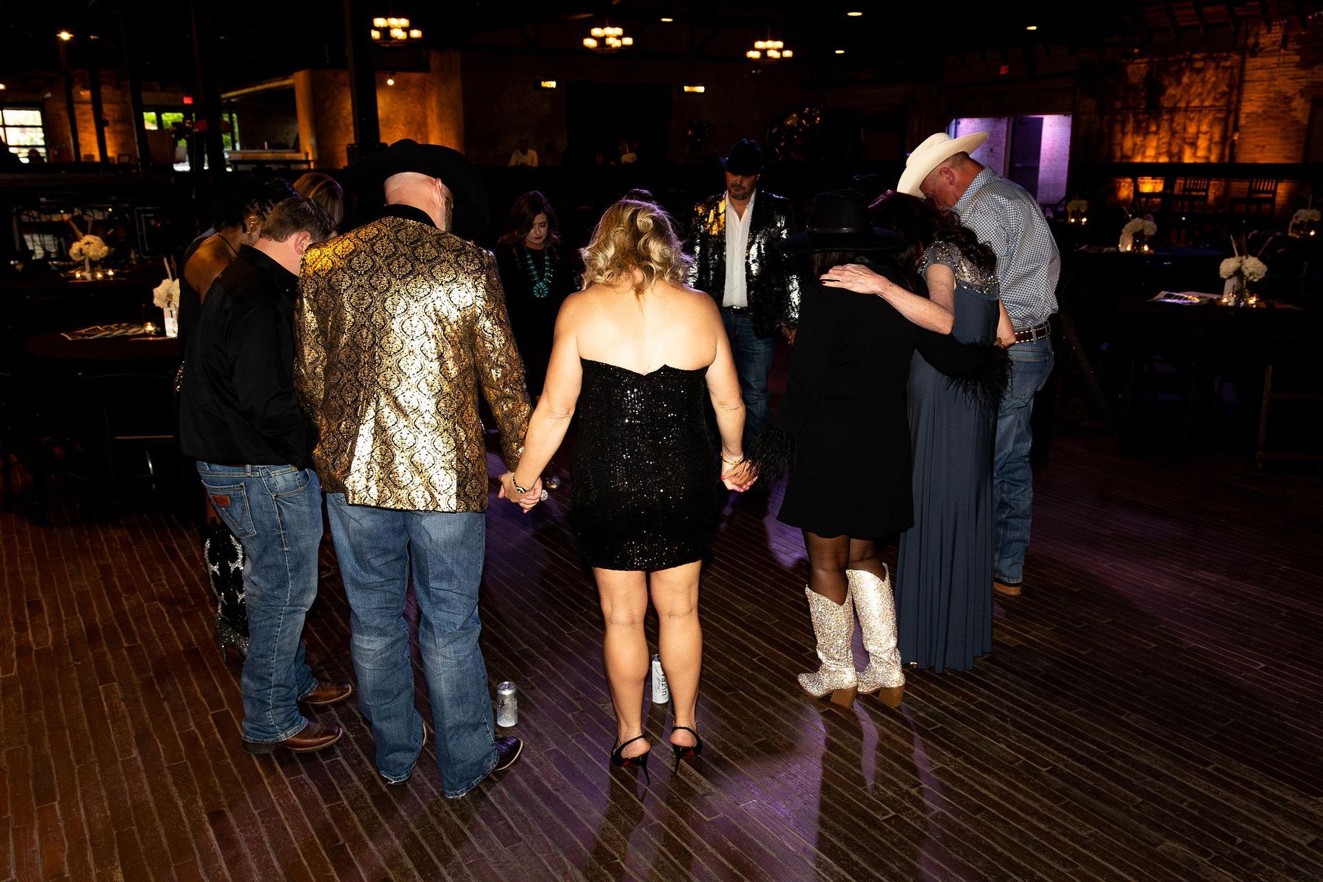 People in a circle dancing at a formal event. Some are wearing cowboy hats and boots. Dark room.