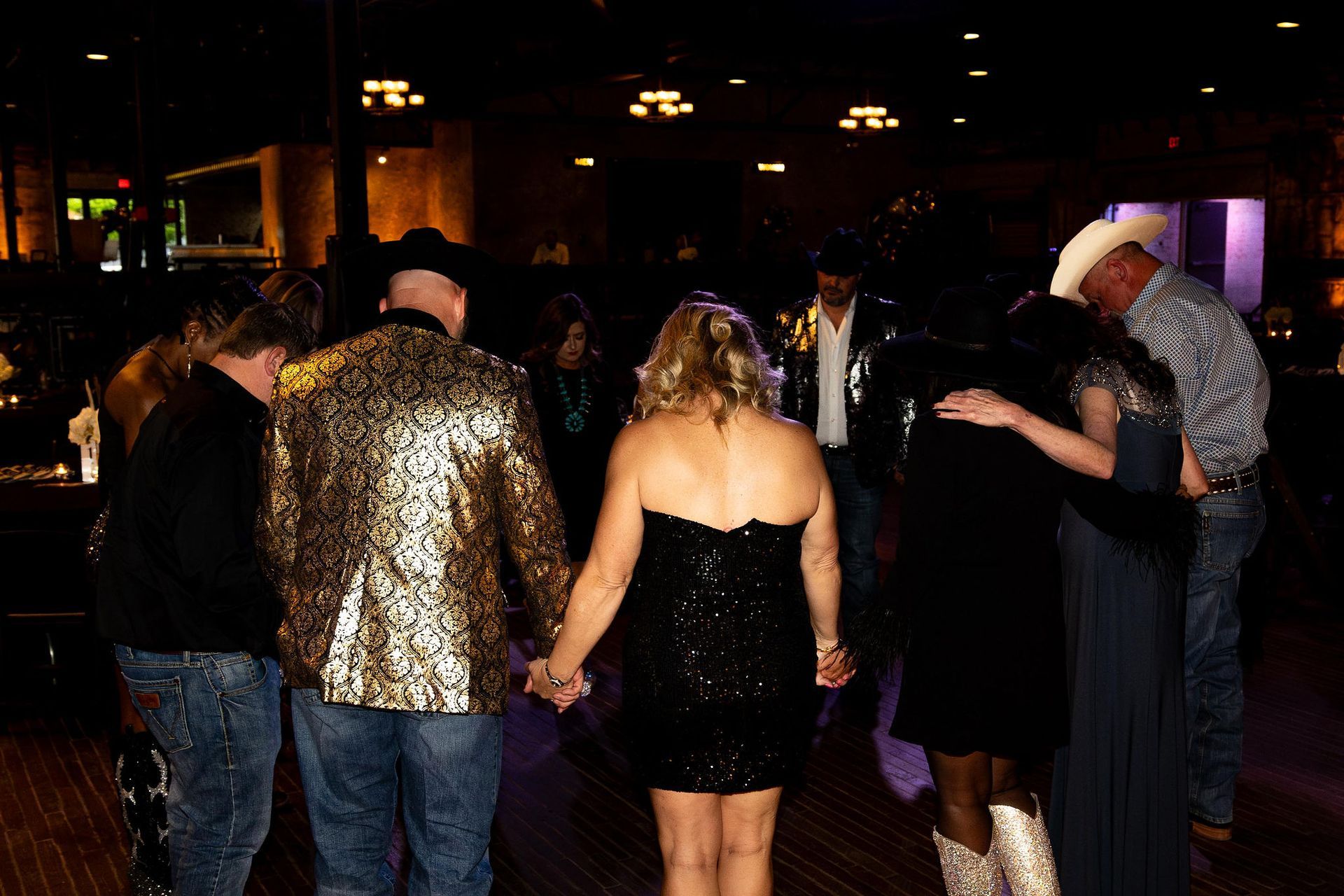 People in formal attire dancing in a dim ballroom, holding hands in a circle.