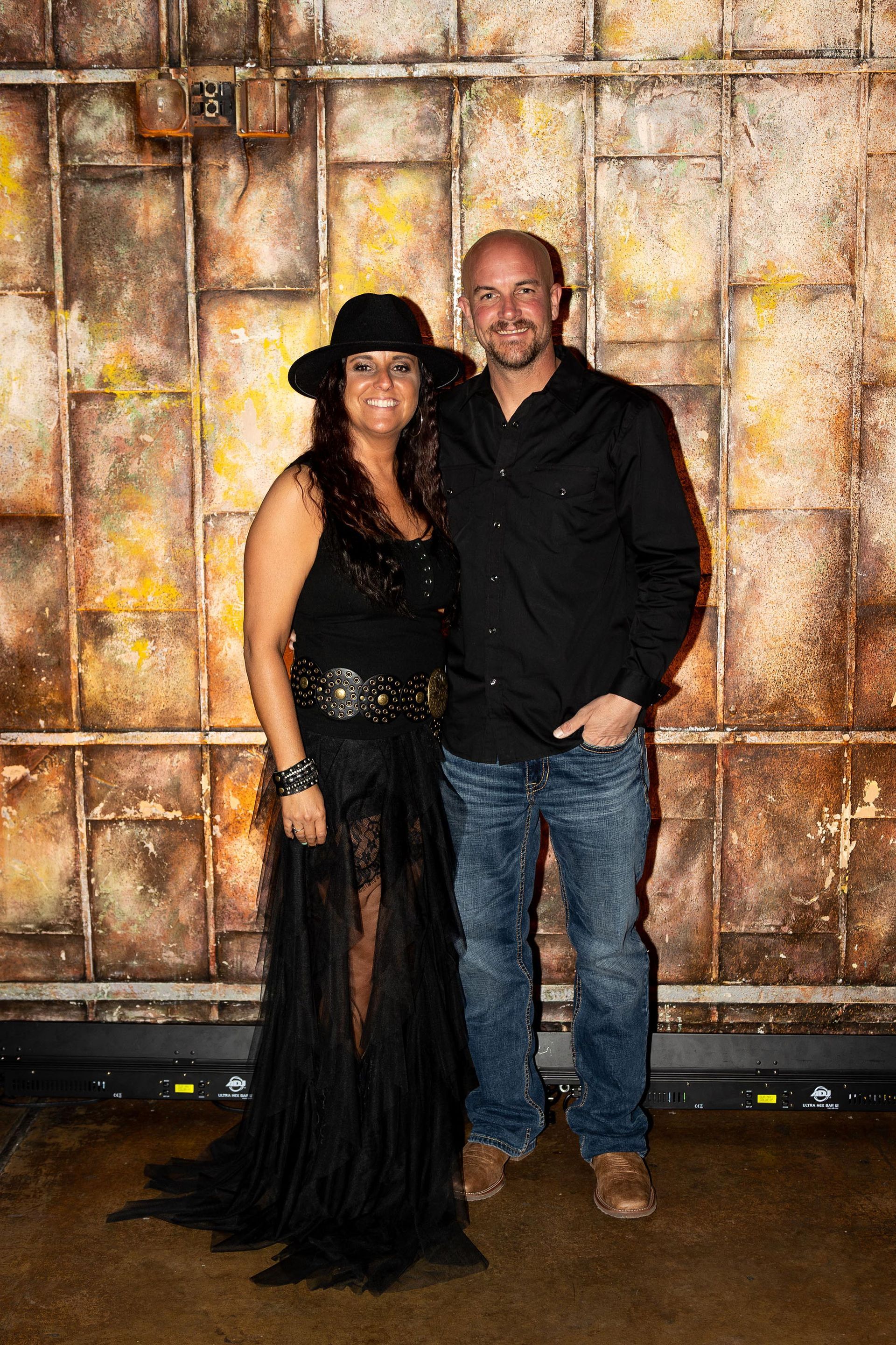 Couple poses in front of a textured wall. Woman in black dress and hat, man in jeans and shirt.