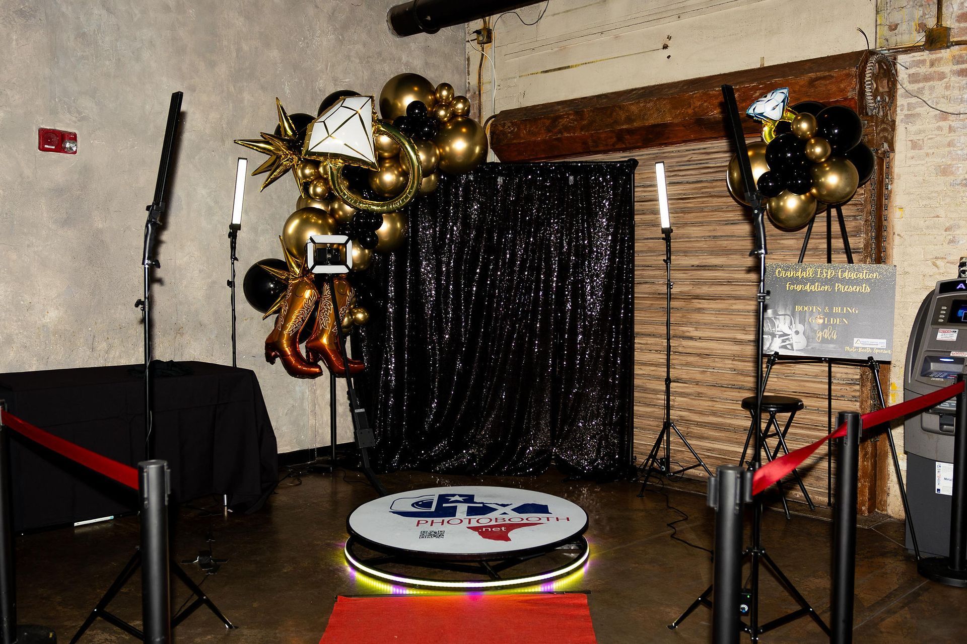 Photo booth setup with a black backdrop, gold and black balloons, and a 360-degree rotating platform.