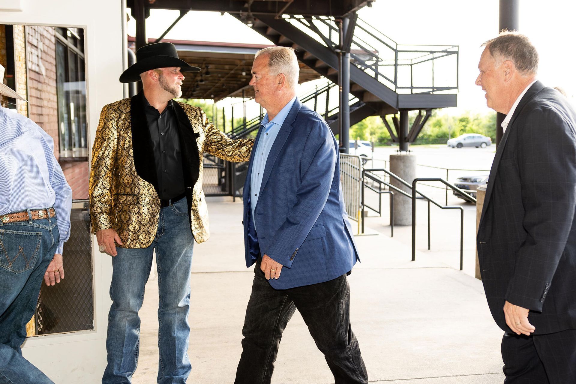 Three men outside, one in gold jacket and cowboy hat, conversing with others in suits.