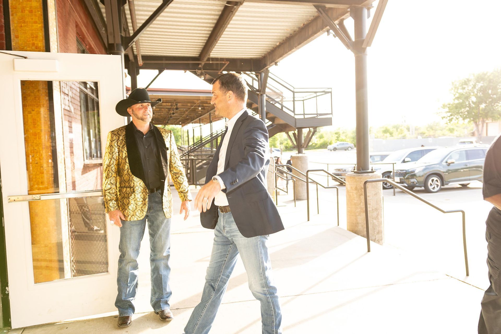 Man in a suit greets another in a sequined jacket and cowboy hat outside a building.