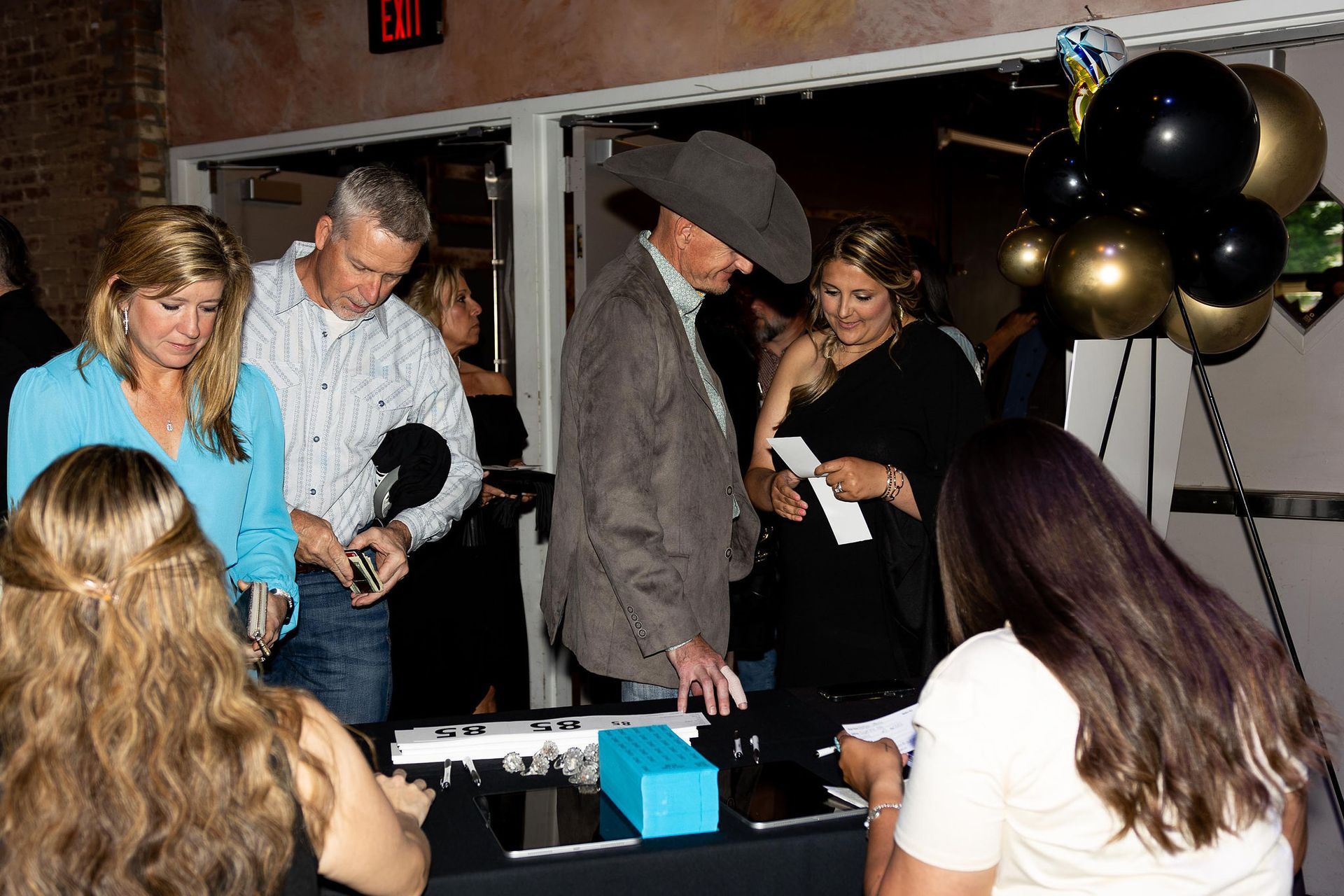 People signing in at an event registration table. Guests include a man in a cowboy hat and balloons in the background.