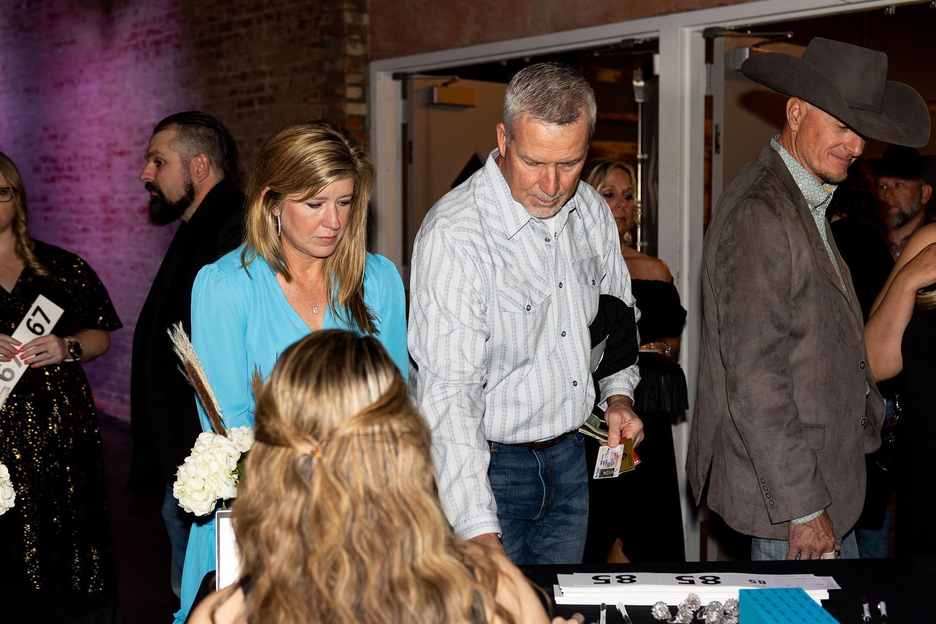 People at an event, registering at a table. Woman in blue dress, man in cowboy hat, others in formal wear.