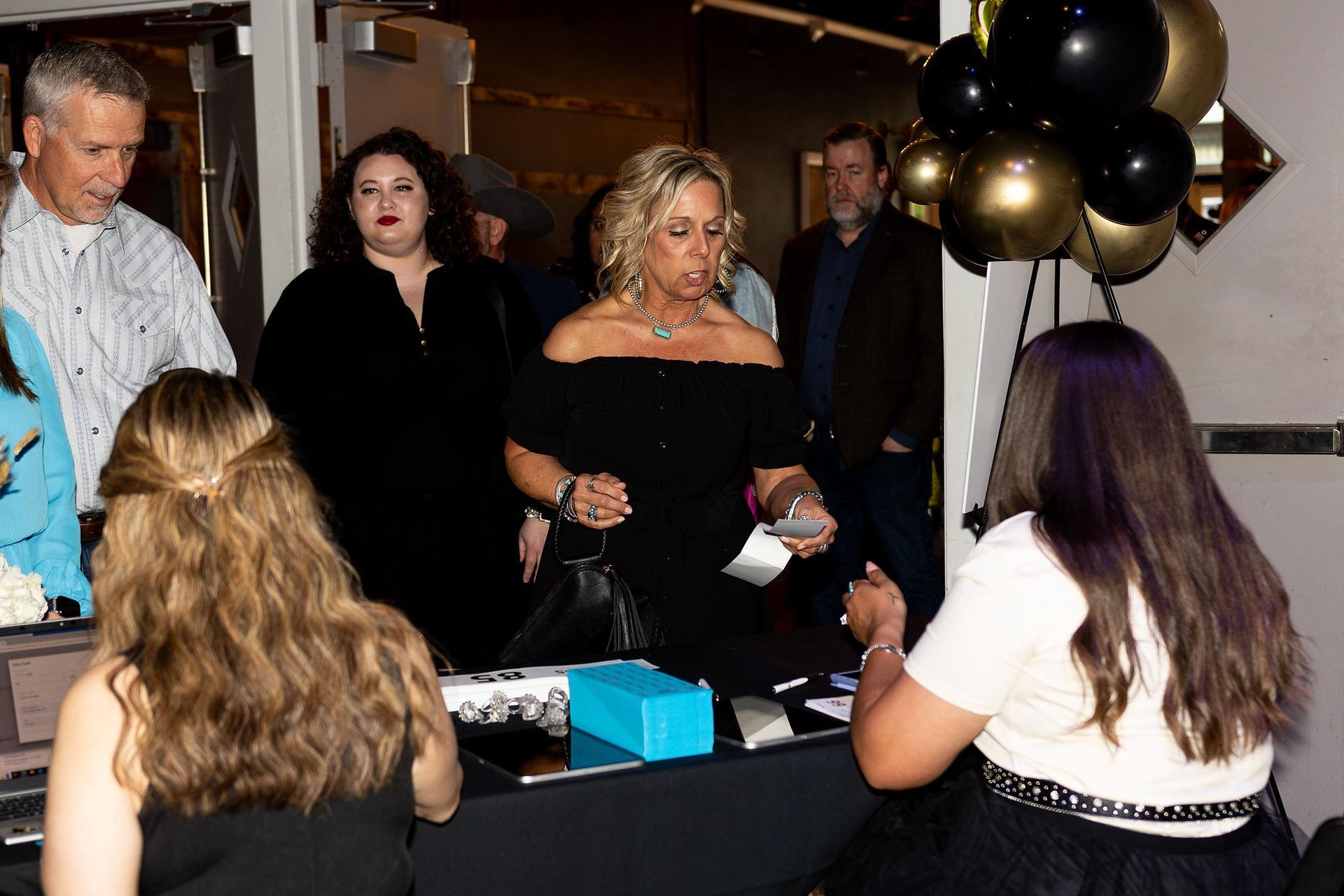 People at a check-in table, a woman in a black dress is being assisted. Balloons in the background.