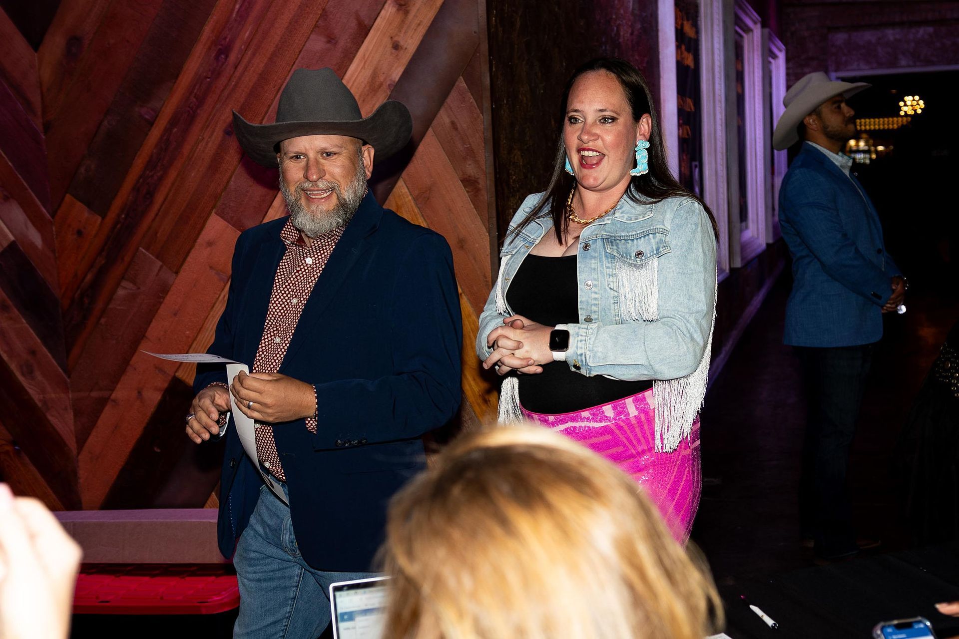 Man and woman on stage with cowboy hats, smiling, holding papers, and a man in the background.