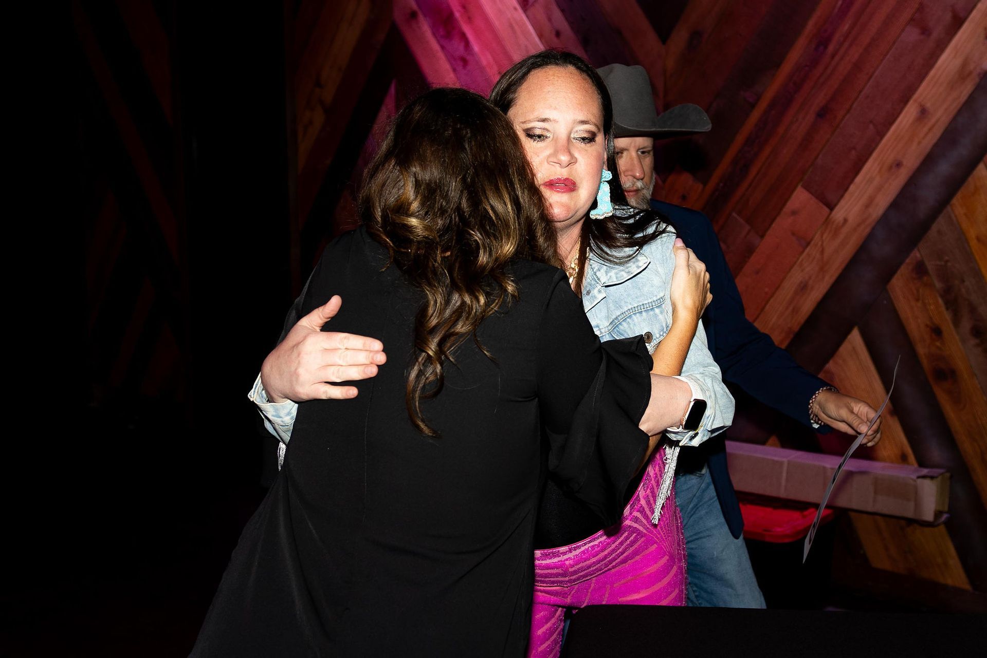 Two women embrace, cowboy looks on with package, wooden backdrop.