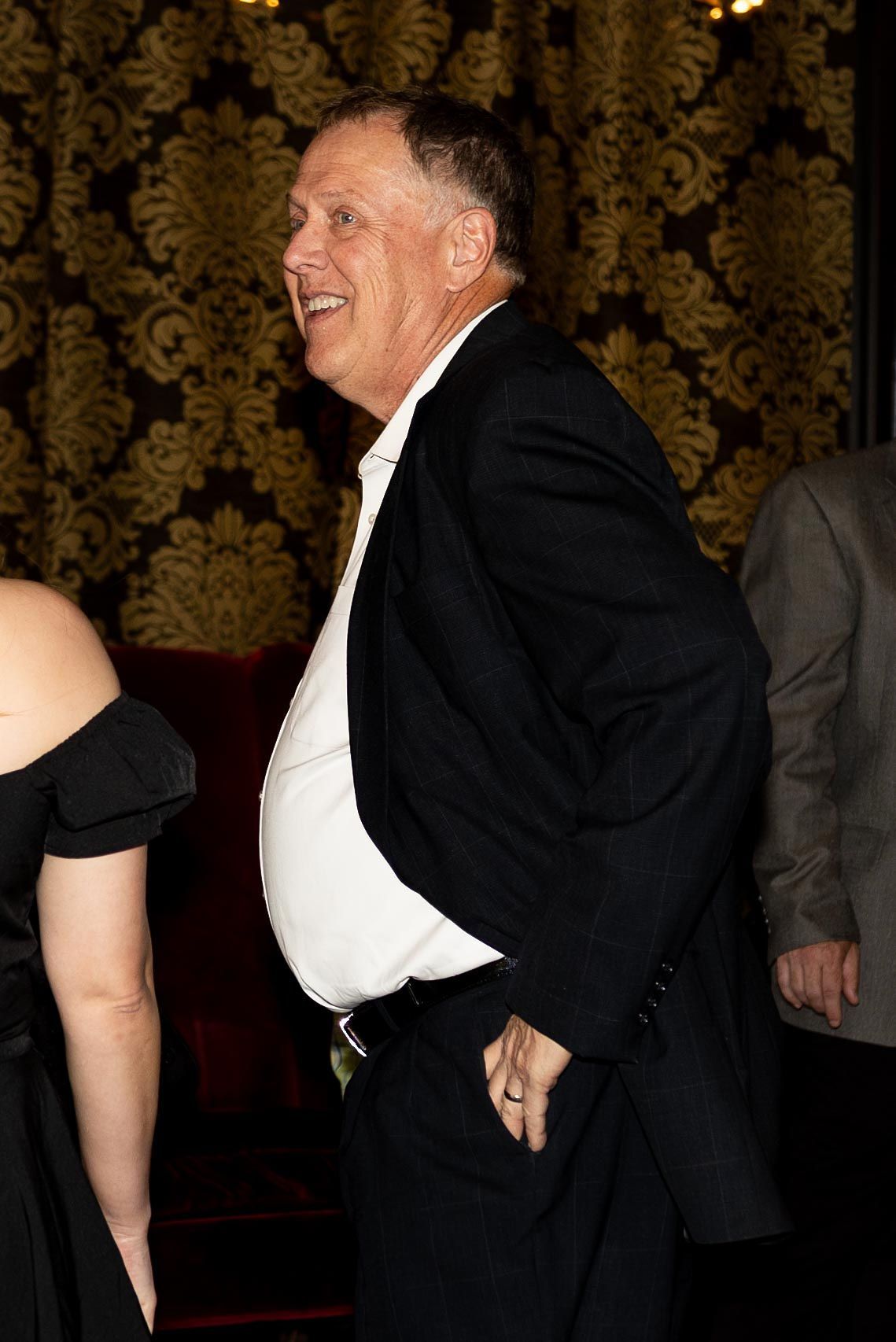 Man in black suit smiles, hands in pockets, inside ornate room with gold wallpaper.