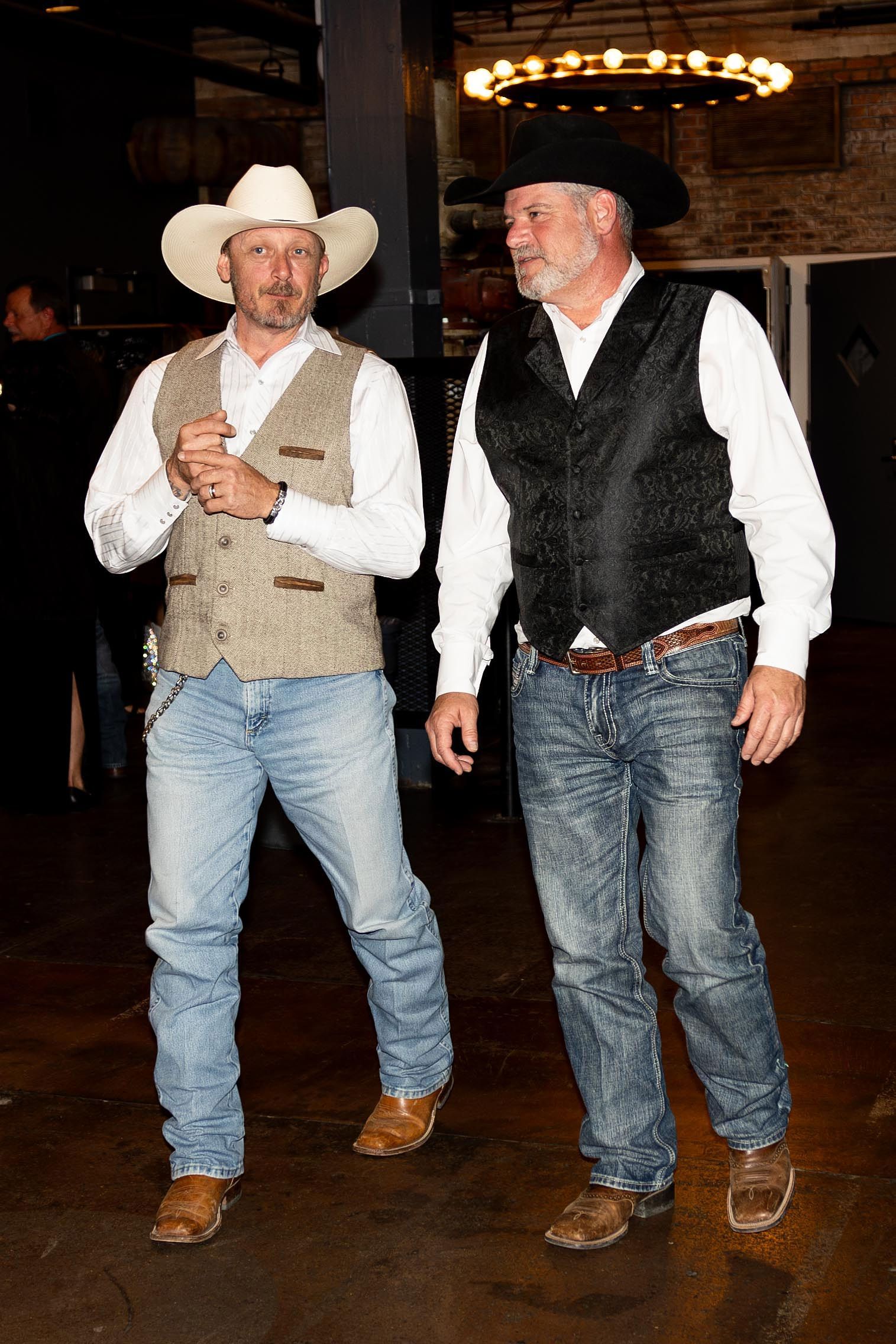 Two men in cowboy attire at an event; one in a tan vest and white hat, the other in a black vest and hat.