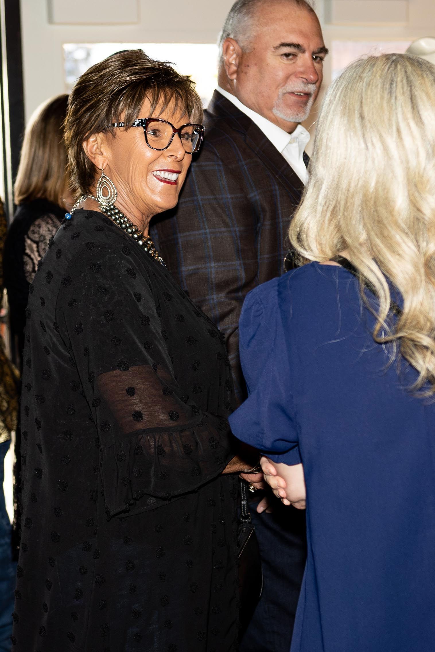 Woman with glasses smiles, speaking to another person. Man in a blazer is behind them.