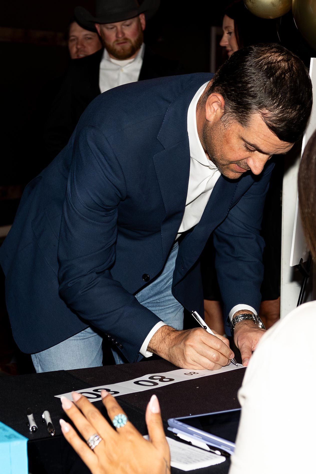 Man in blue suit signs an autograph on a table; person with manicured nails holds a pen in the foreground.