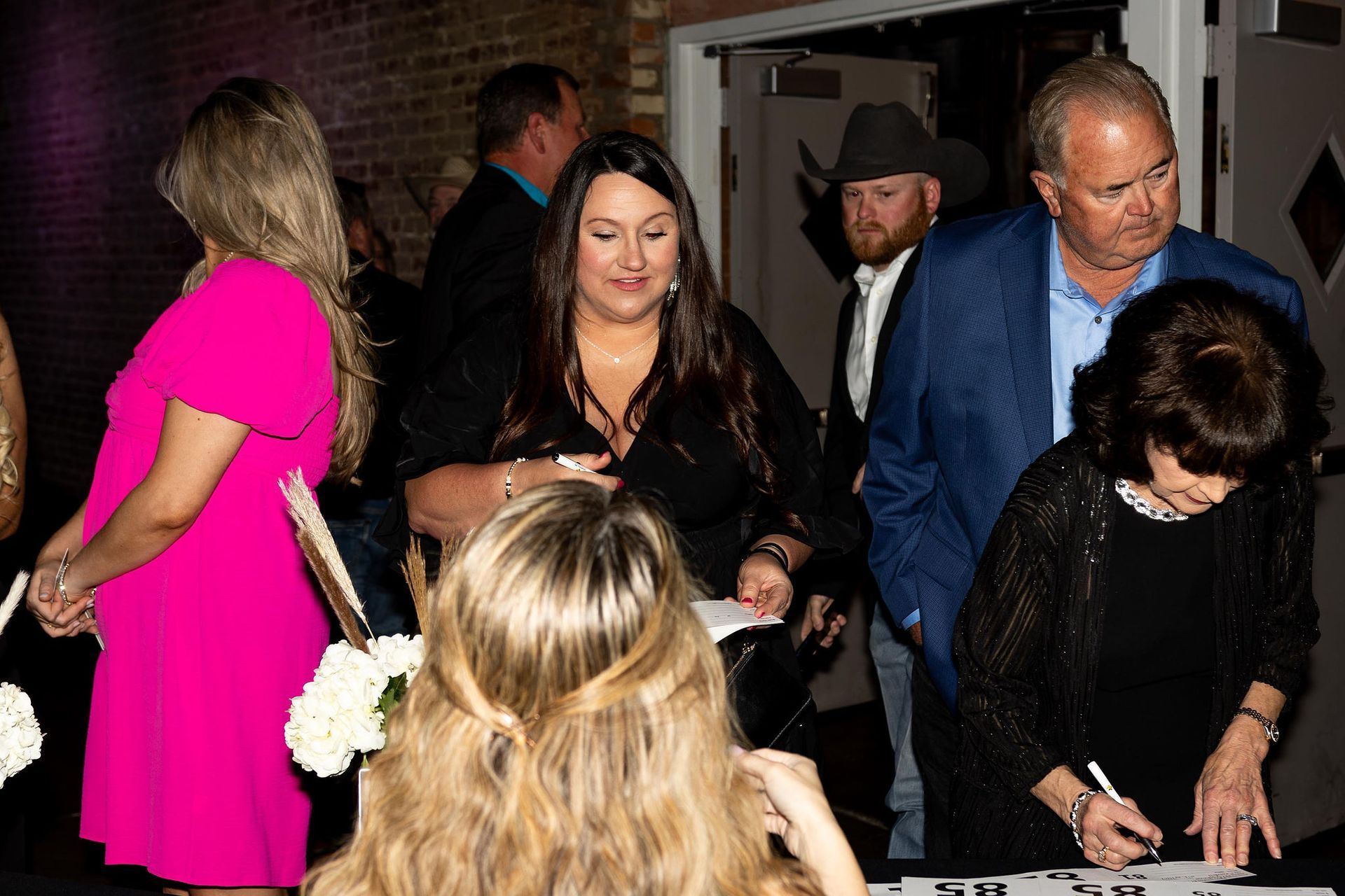 People signing a guest book at an event. Woman in pink dress, others in various attire. Indoor setting.